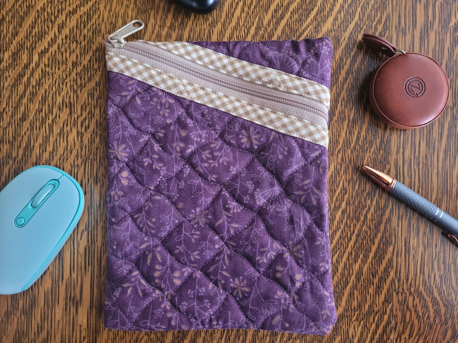 Purple quilted pouch with a zipper on a wooden surface with a mouse, pen, and case.