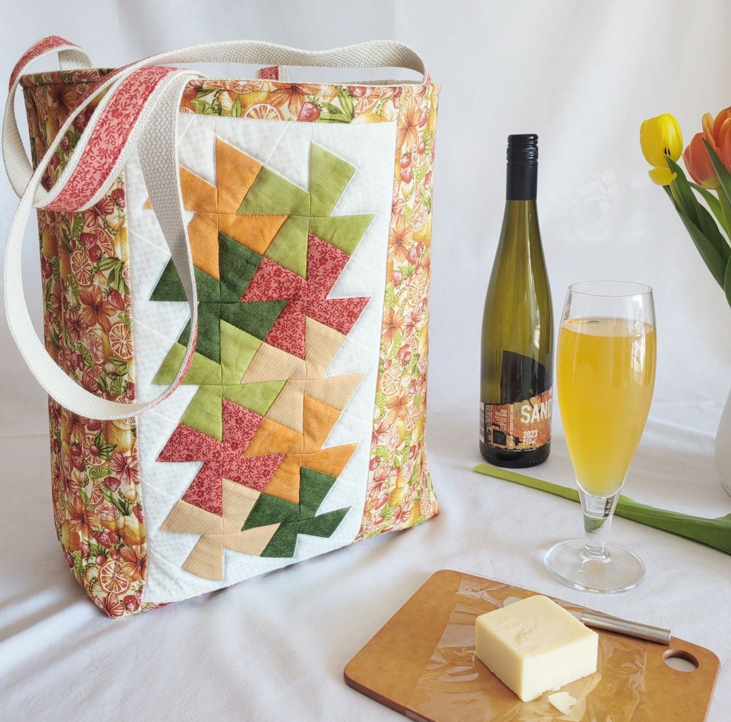 Colorful quilted bag with a bottle of wine, glass, and cutting board on a white surface