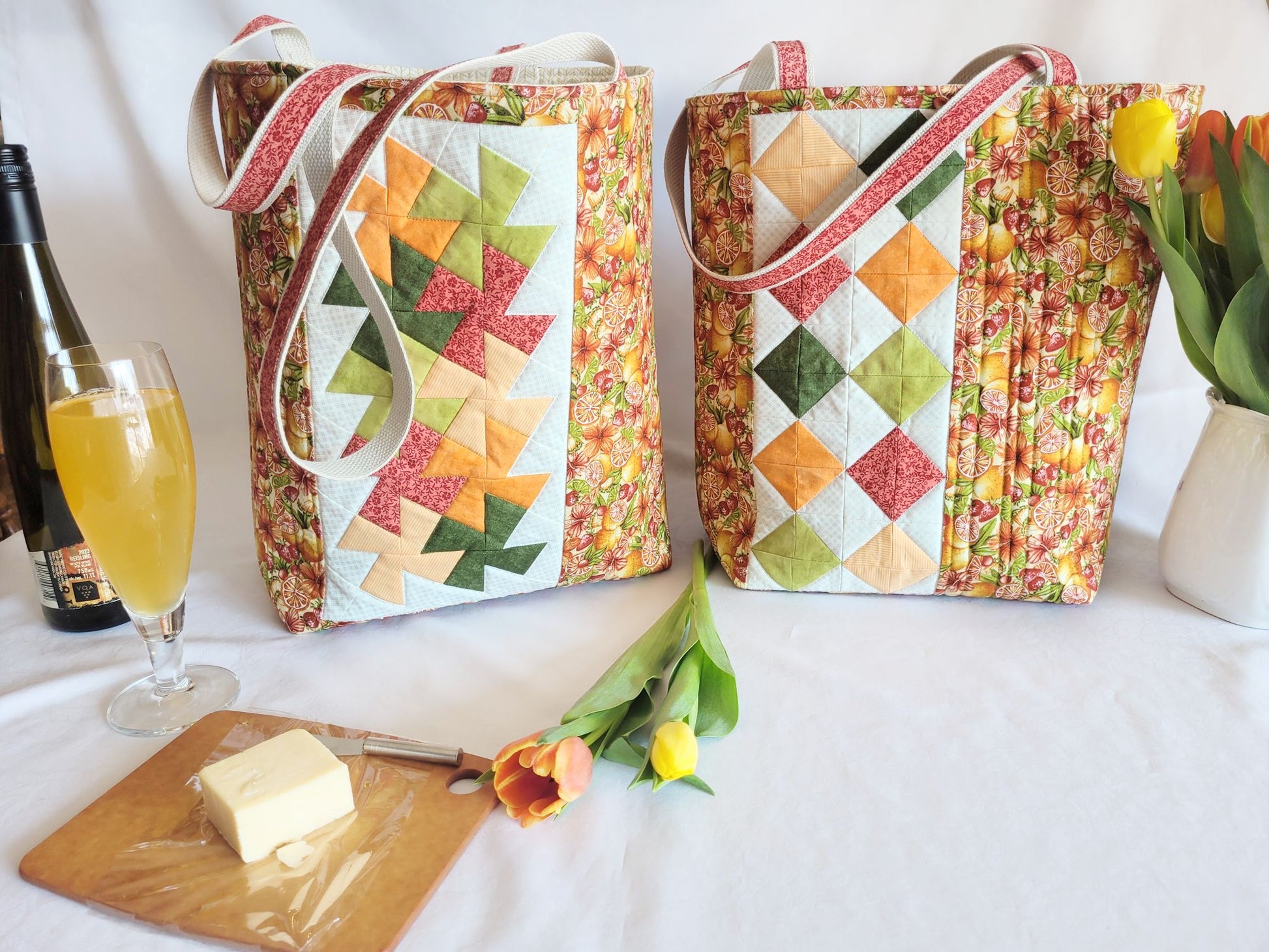 Two colourful quilted bags on a table with a glass of cider, bottle, and flowers.