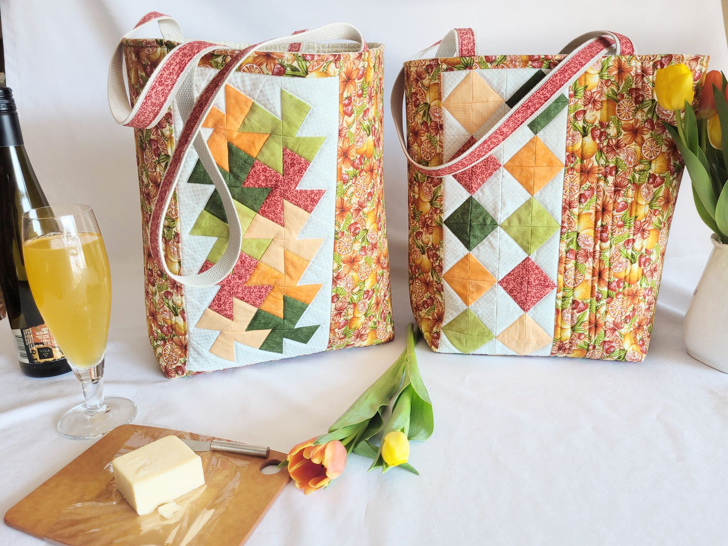 Two quilted bags with a glass of cider, bottle, and flowers on a white surface.