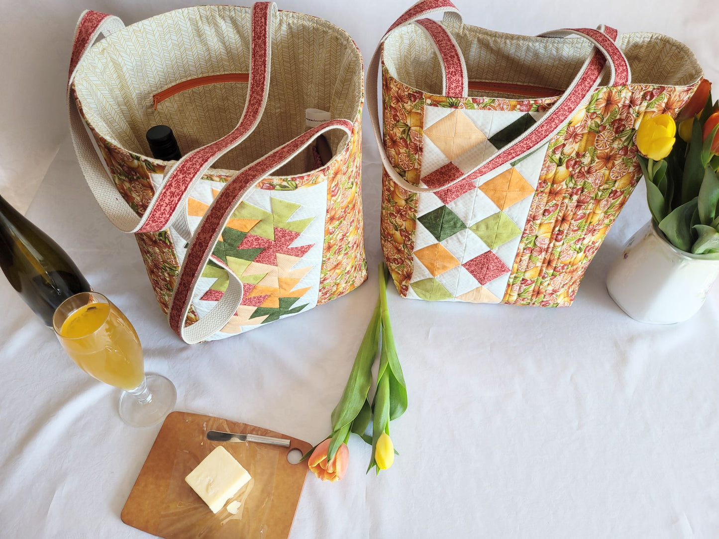 Two fabric bags with wine bottles on a table with a glass of juice, cutting board, and tulips.