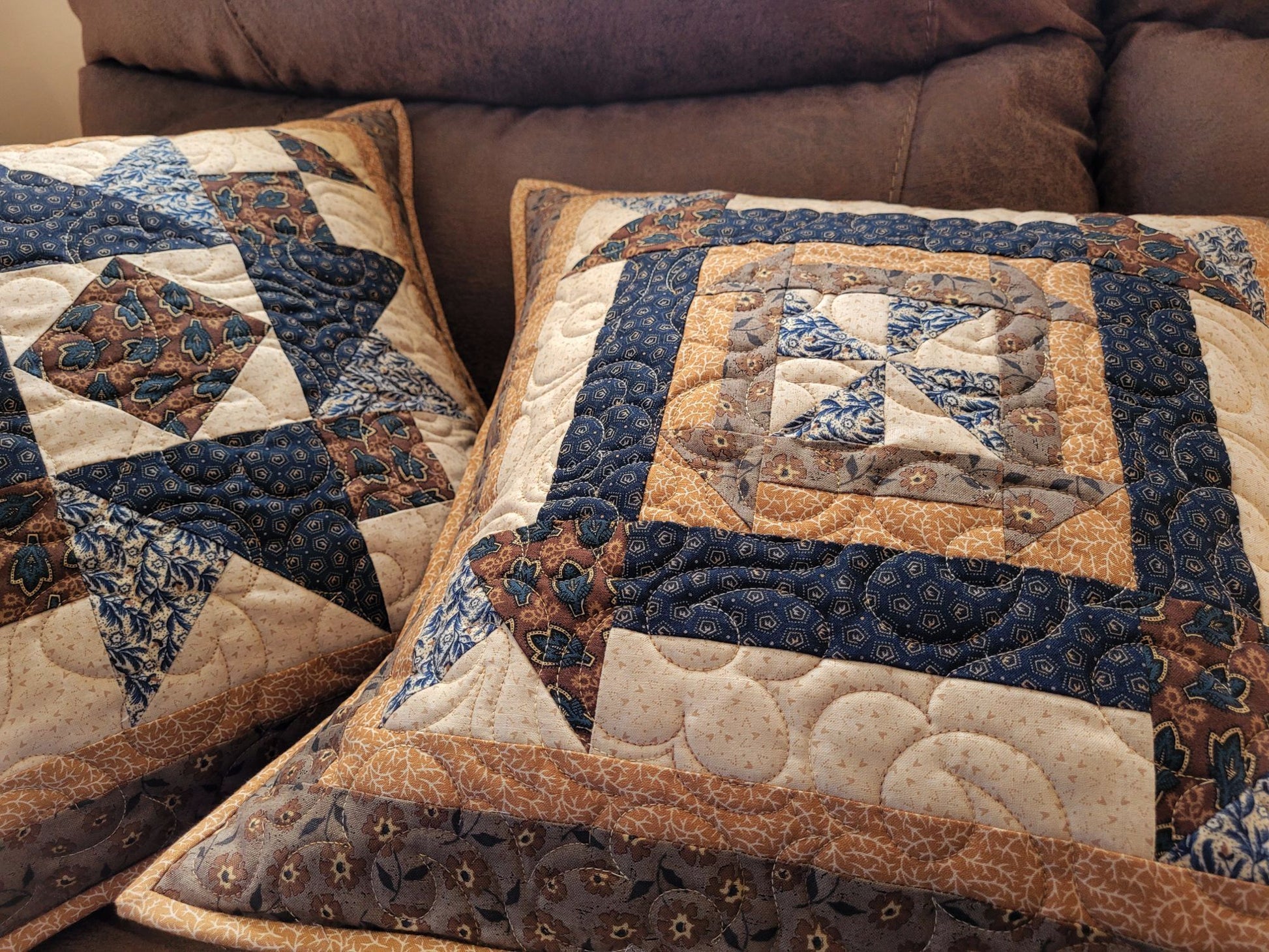 Quilted pillows with a patchwork design on a brown sofa in an angled view to show quilting texture.