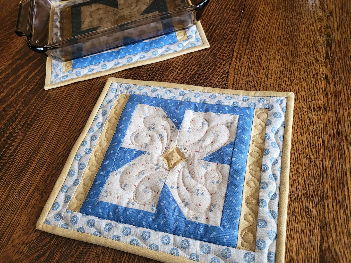 Large quilted hot mats on a wooden table with one holding an 11 x 9 inch casserole in the background.