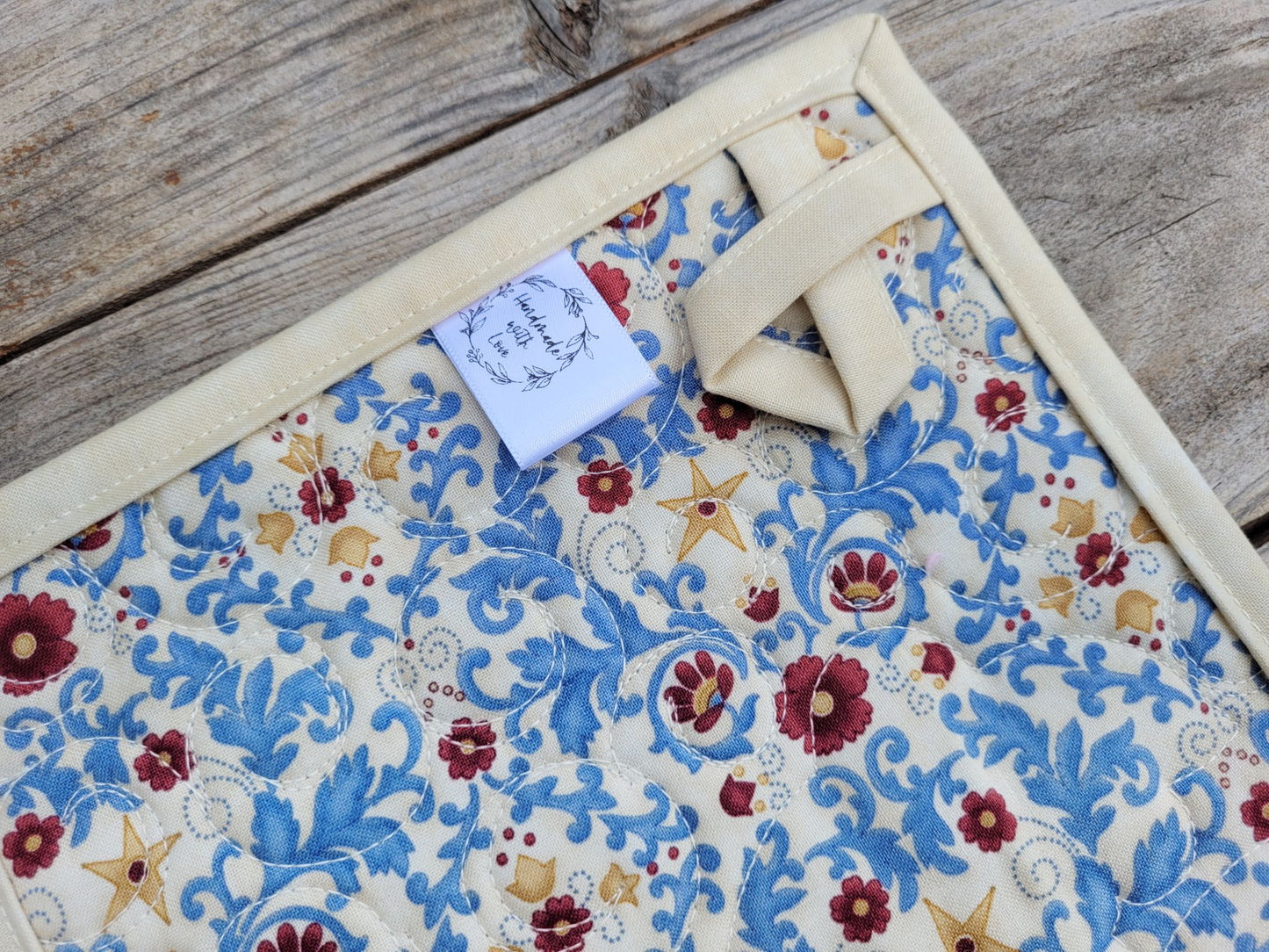 Back of quilted potholder showing hanging loop, handmade label and floral fabric, on a wooden surface. 