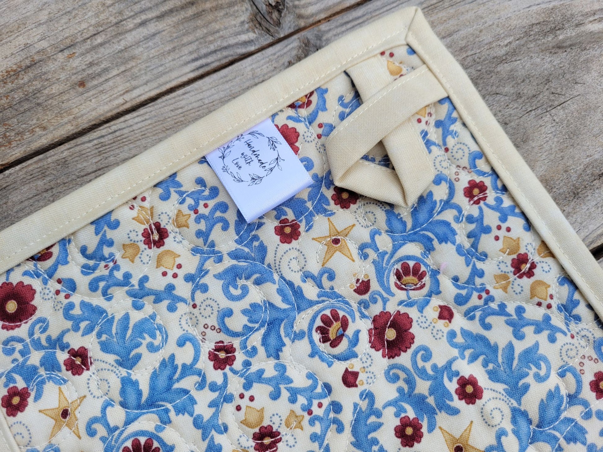 Back of quilted potholder showing hanging loop, handmade label and floral fabric, on a wooden surface. 