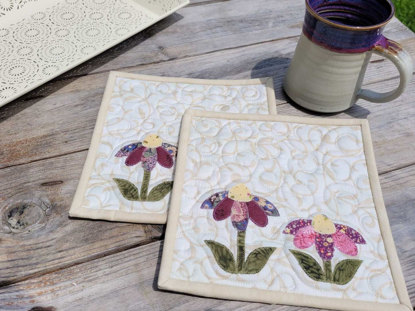 Two floral hot mats on a wooden surface with a mug.