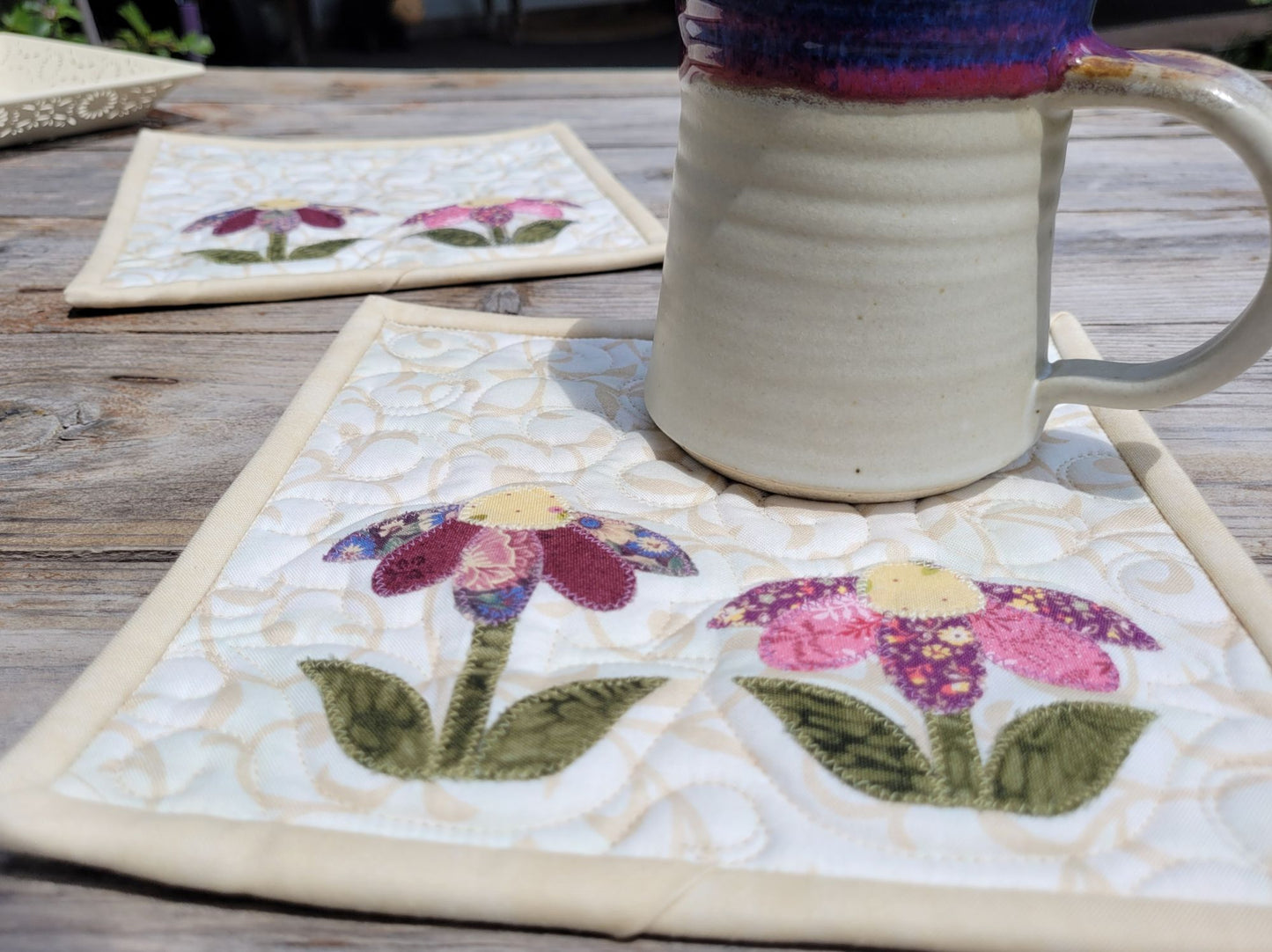 Two quilted flower potholders with a pottery mug on an outdoor wood table.