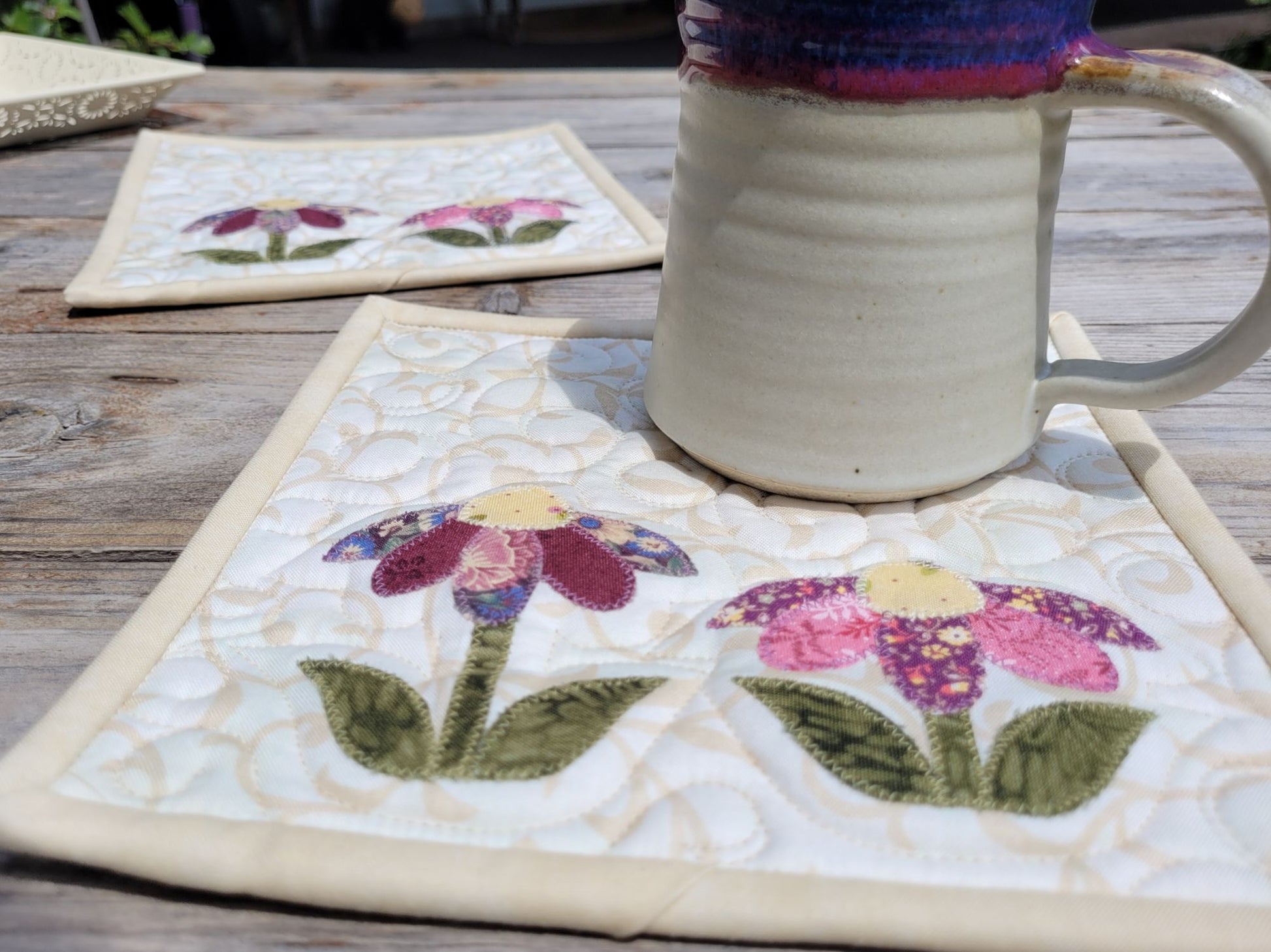 Two quilted flower potholders with a pottery mug on an outdoor wood table.