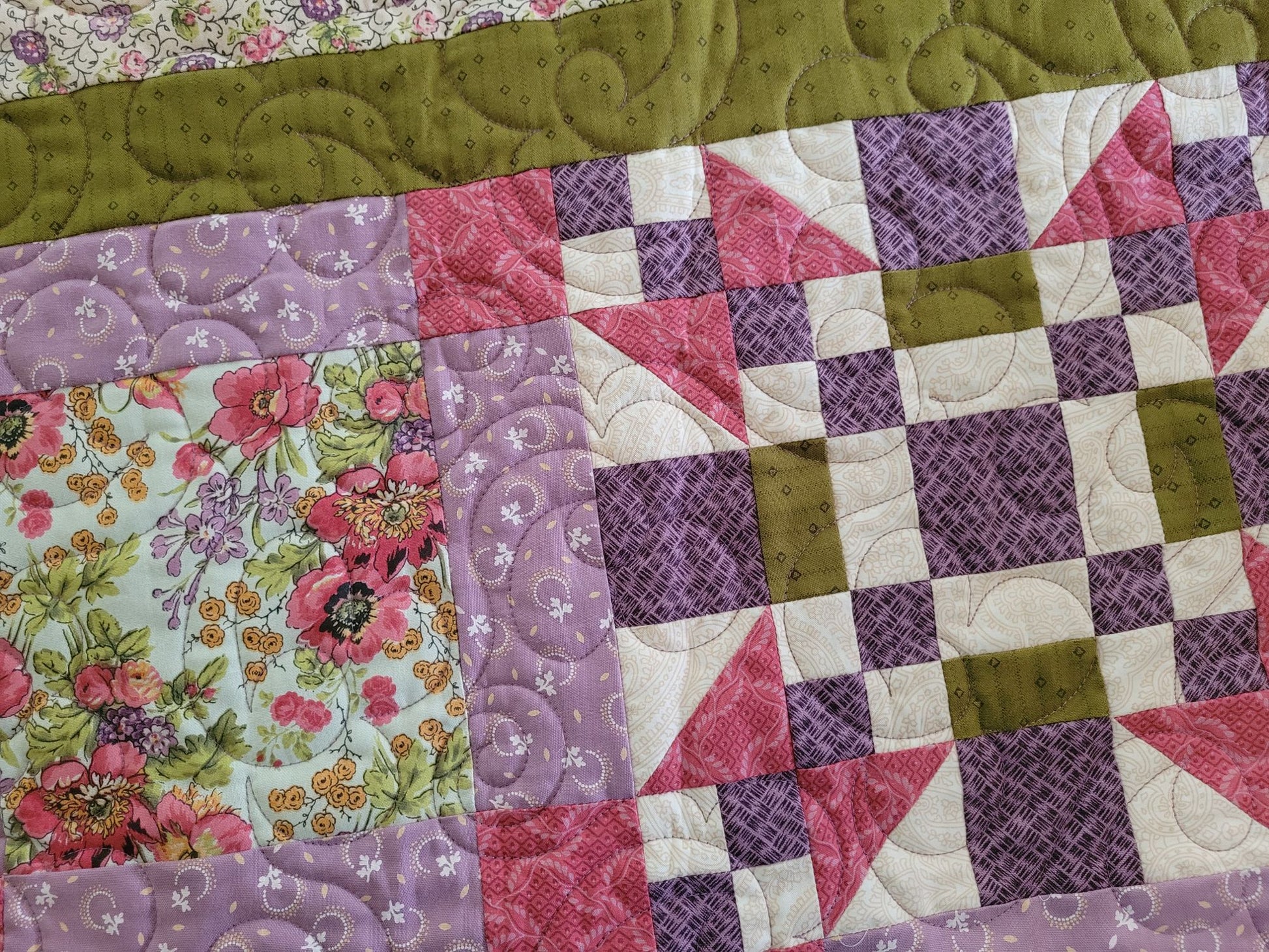 A close-up of a handmade quilt featuring a patchwork design with purple floral prints, pink and white geometric blocks, and an moss green border.