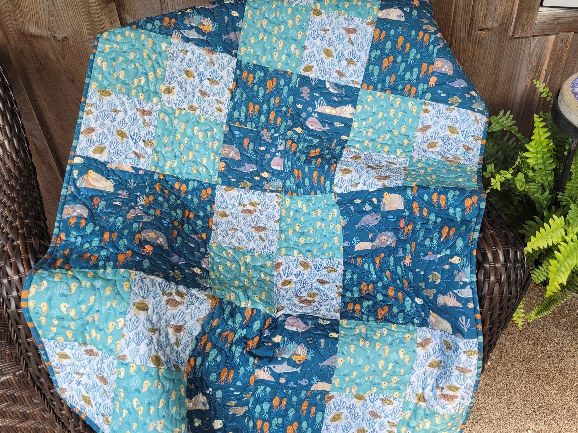 A handmade patchwork quilt featuring ocean-themed prints of whales, fish, and seashells in shades of blue, teal, and white, draped over a dark brown wicker chair next to a potted green fern.