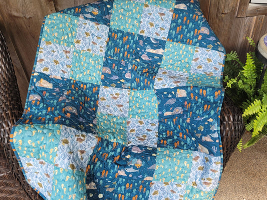 A handmade patchwork quilt featuring ocean-themed prints of whales, fish, and seashells in shades of blue, teal, and white, draped over a dark brown wicker chair next to a potted green fern.