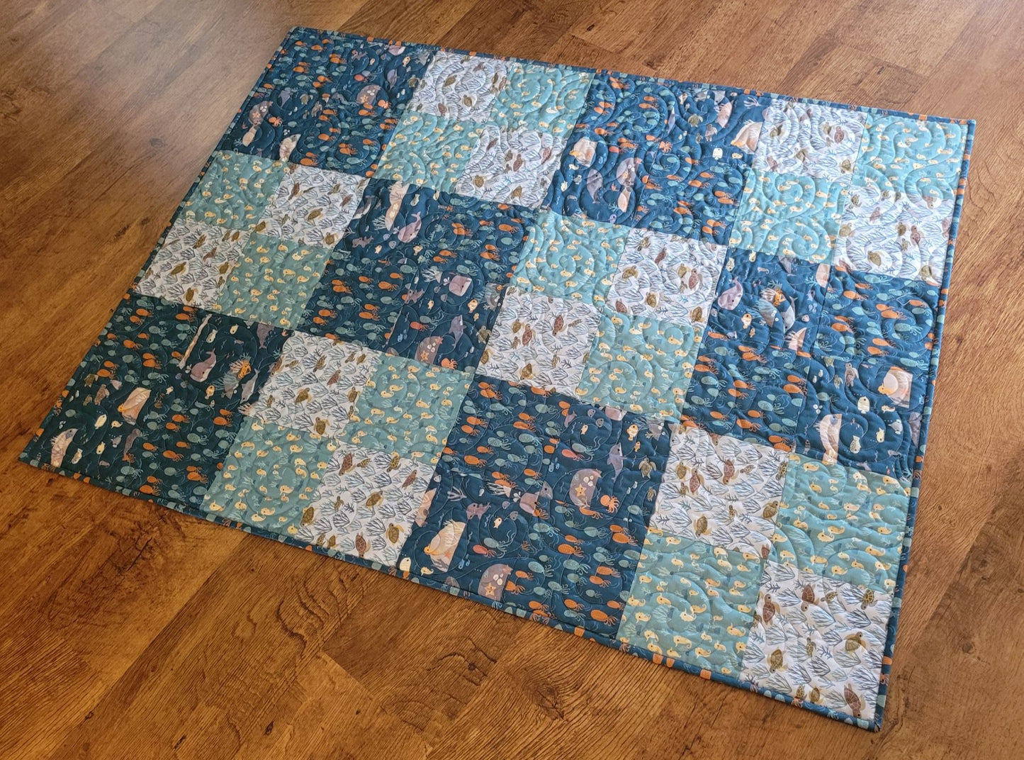 A handmade patchwork quilt with a blue and teal colour scheme, featuring various ocean and sea animals and nature-themed prints, laid out on a wooden floor.
