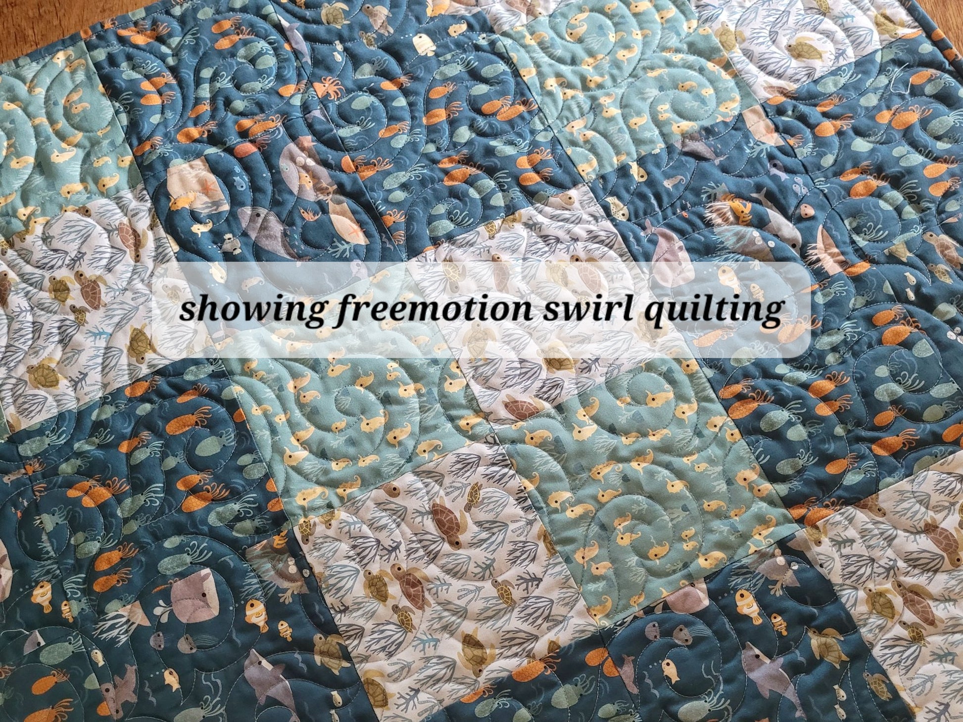 A close-up of a baby quilt with patchwork four patch squares in blue, teal, cream, and orange-brown tones, featuring swirling stitched patterns and overlaid text reading 'showing freemotion swirl quilting'.