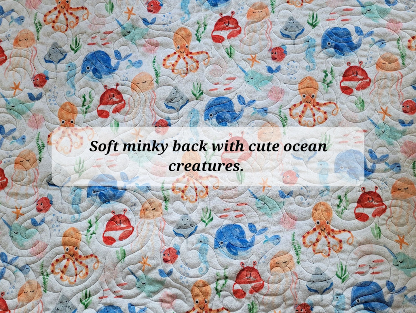 A close-up of a quilted fabric with a colourful ocean-themed print, including blue whales, red crabs, orange octopuses, and sea plants on a light background. A semi-transparent text box in the centre reads, 'Soft minky back with cute ocean creatures'.