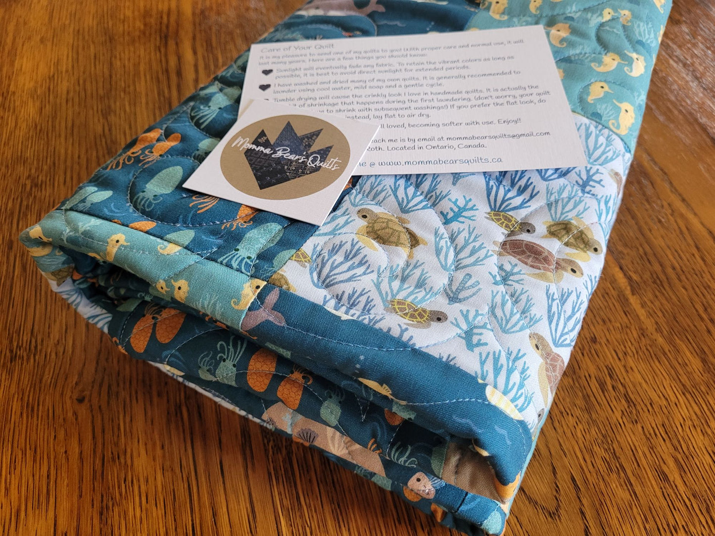 A folded quilted baby blanket with a blue and white sea life pattern, featuring turtles, dolphins, and coral, resting on a wooden table. A circular 'Momma Bears Quilts' brand business card and a printed care/instruction card are placed on top.