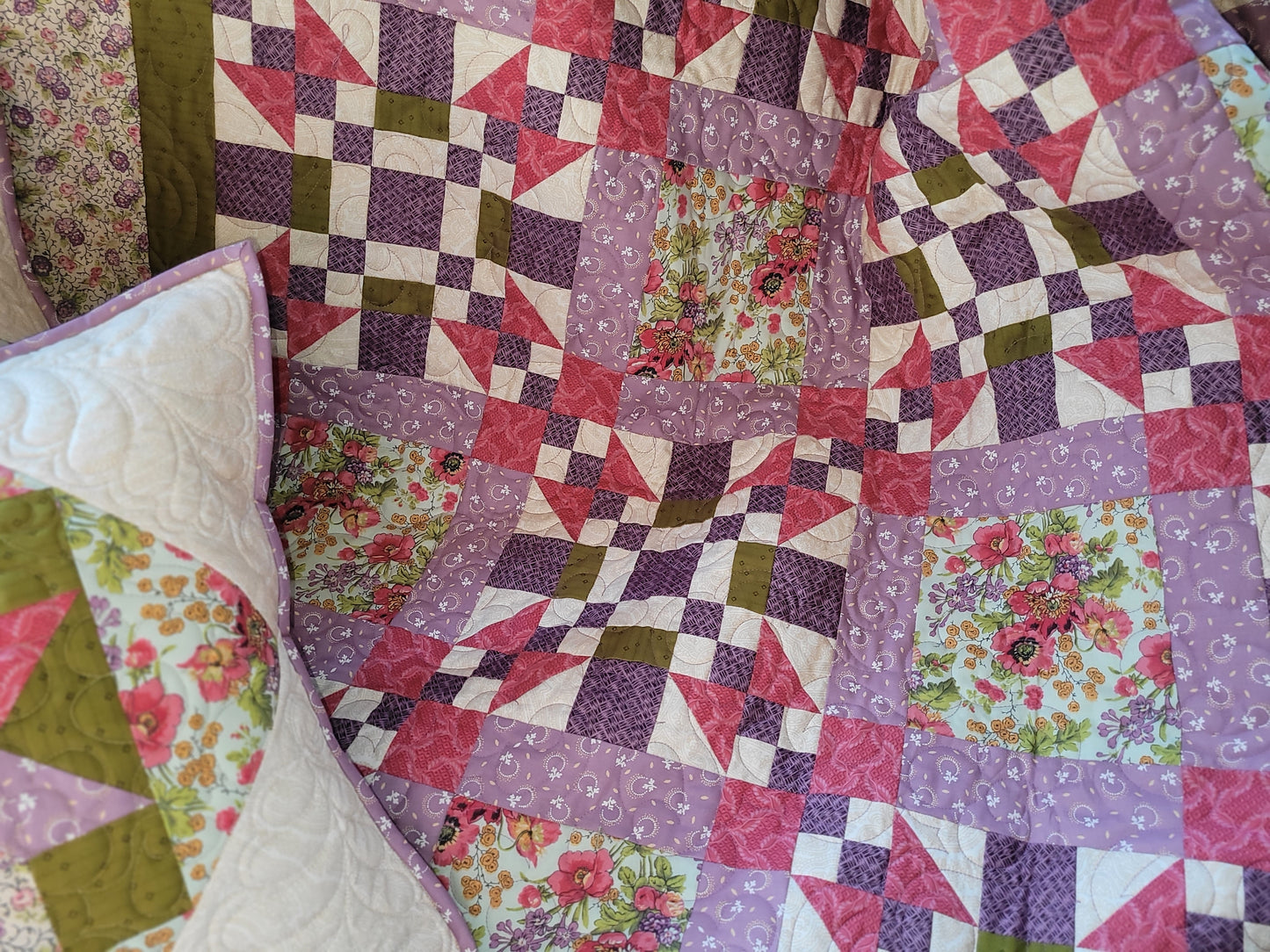 A close-up of a handmade quilted blanket featuring a patchwork design of purple, pink, and moss green geometric and floral patterns.