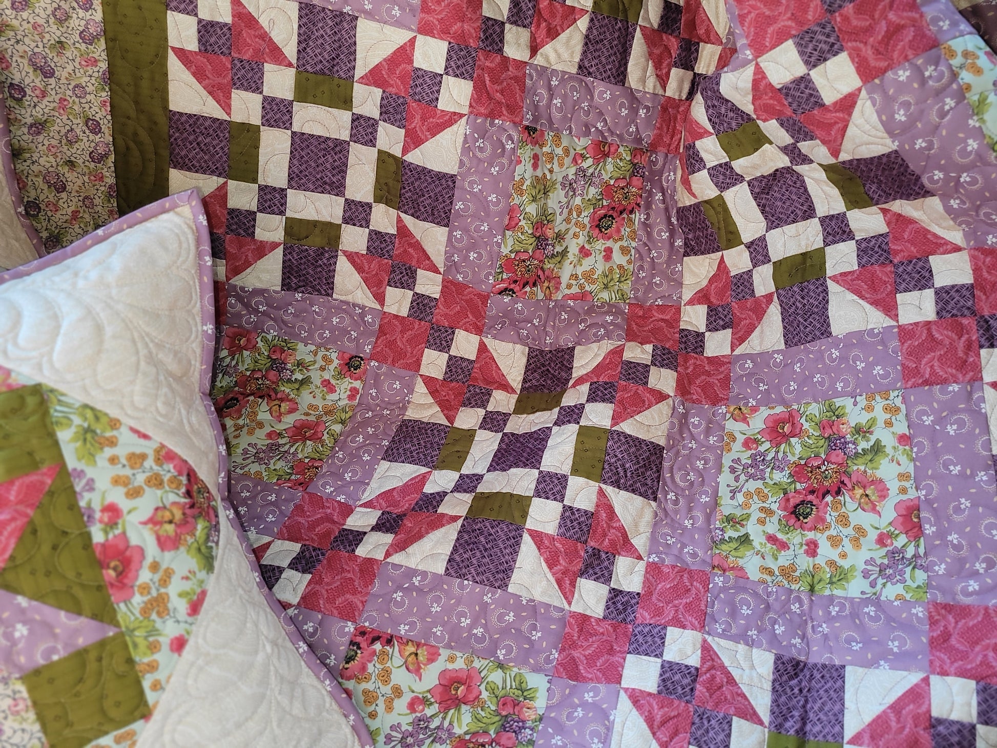 A close-up of a handmade quilted blanket featuring a patchwork design of purple, pink, and moss green geometric and floral patterns.