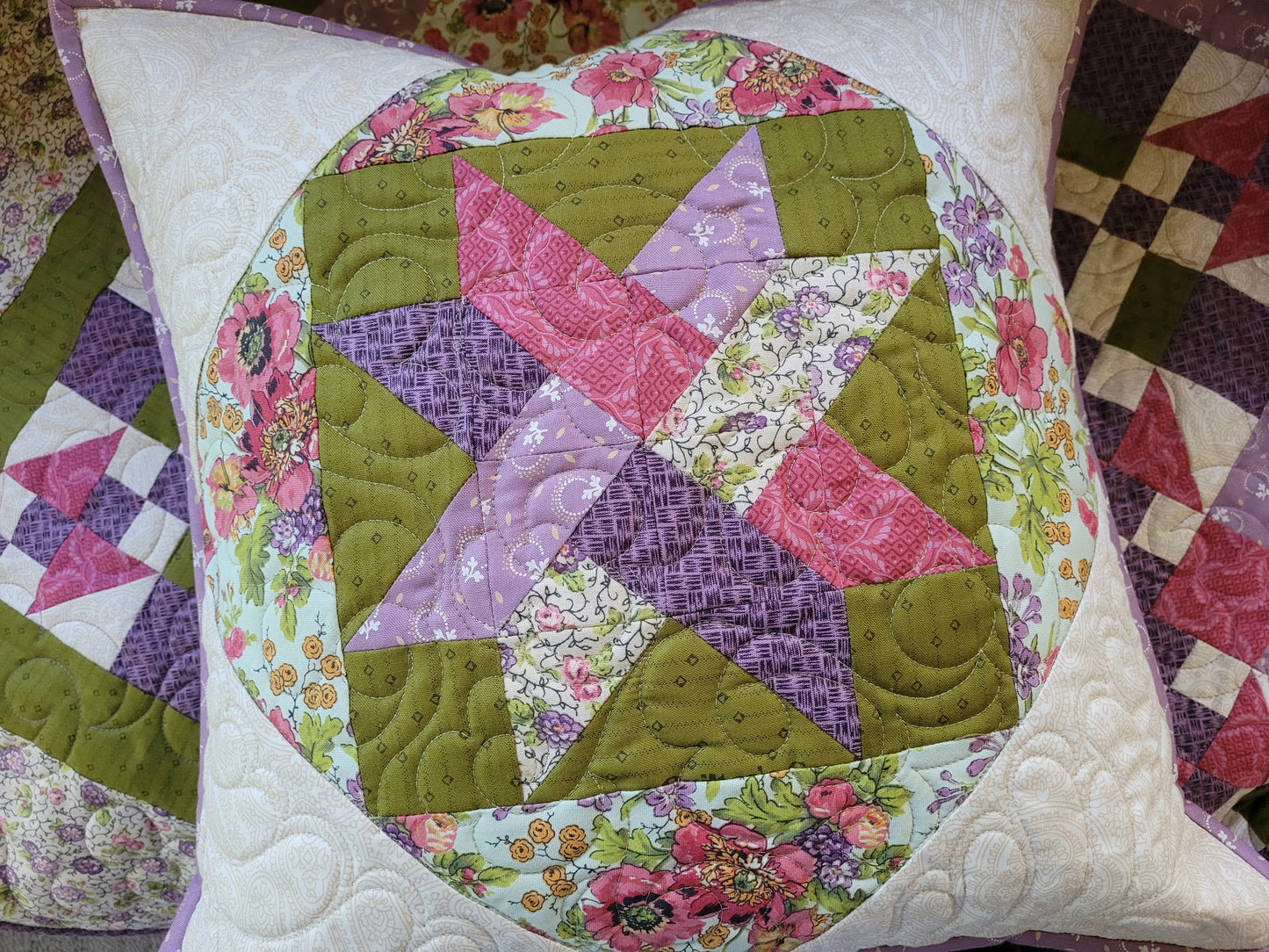 A square quilted throw pillow featuring a colorful patchwork starburst design on moss green with pink, purple, and floral patterned sections on a white quilted border.