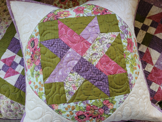 A square quilted throw pillow featuring a colorful patchwork starburst design on moss green with pink, purple, and floral patterned sections on a white quilted border.