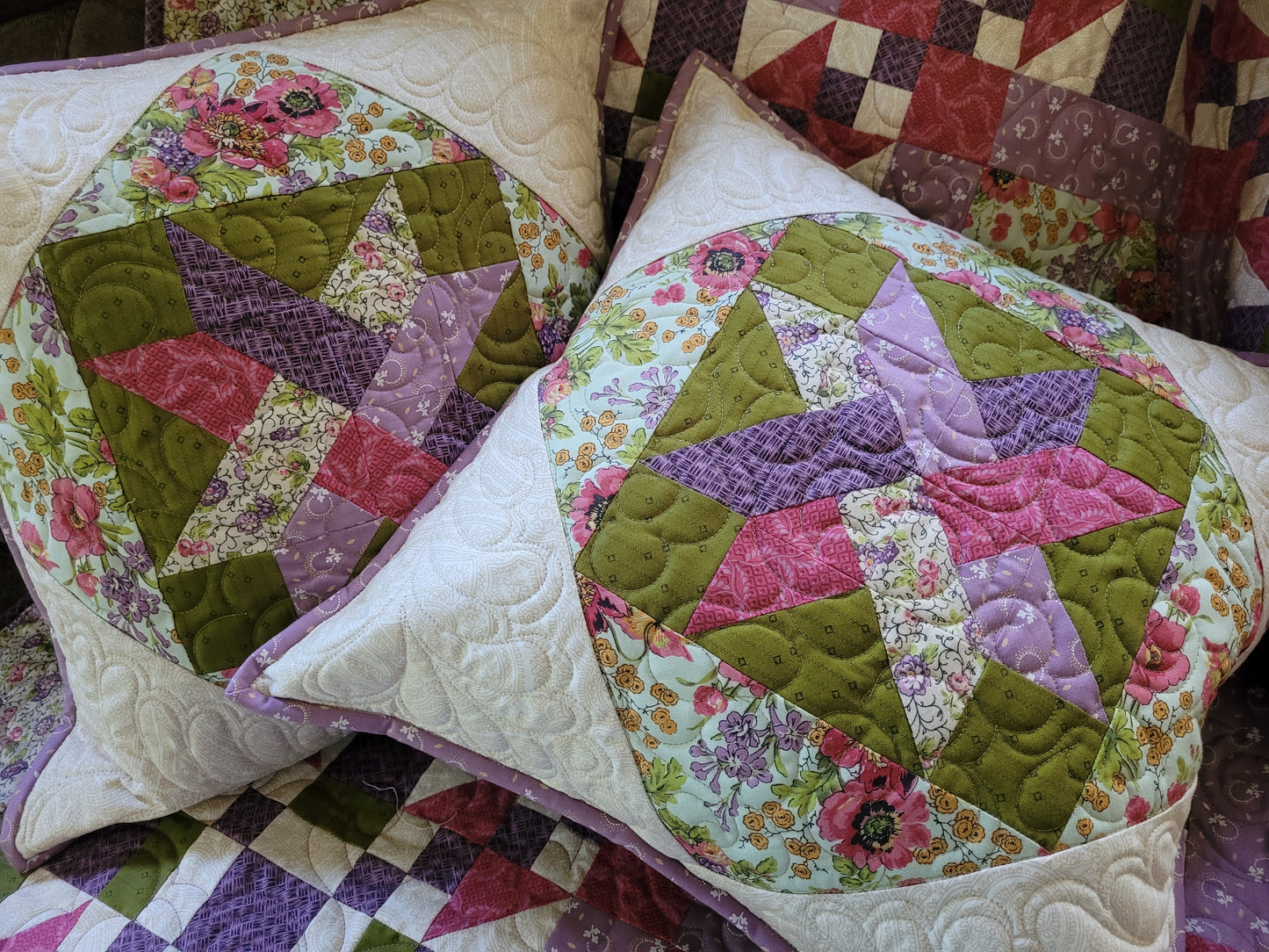 Two square quilted throw pillows with circular starburst patchwork designs in moss green, purple, pink, and floral prints, resting on a matching quilted blanket.