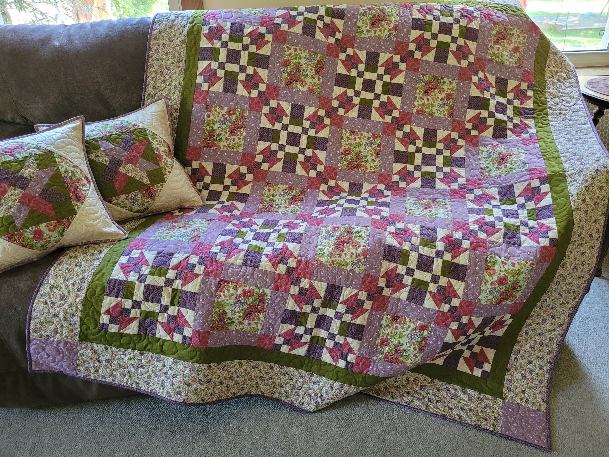 A multicolor patchwork quilt with floral and geometric patterns draped over a dark brown leather couch, accompanied by two matching decorative throw pillows.