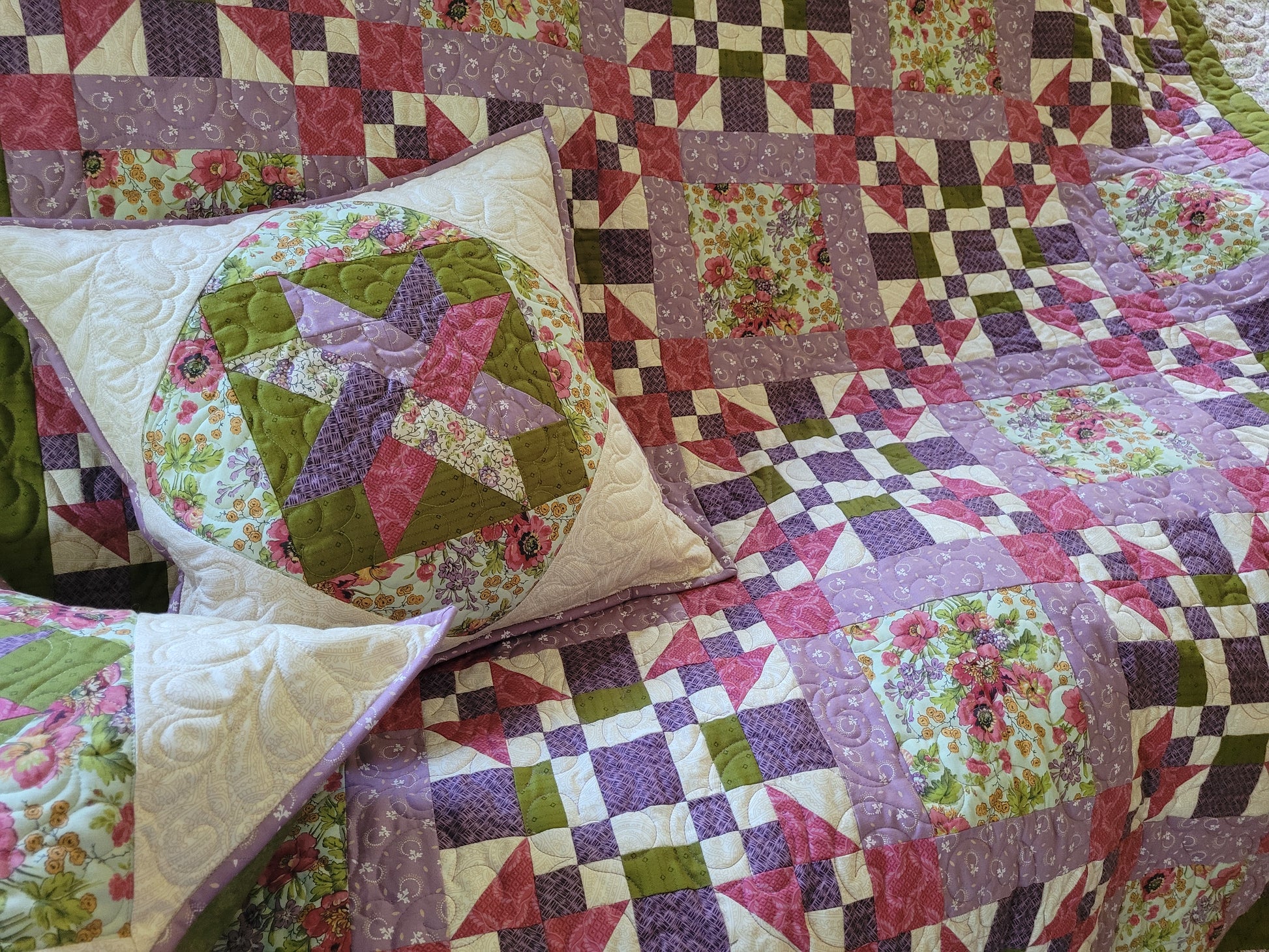 A colorful patchwork quilted blanket and matching pillows displayed on a sofa. The quilt features a mix of purple, green, pink, and cream floral and geometric patterns.