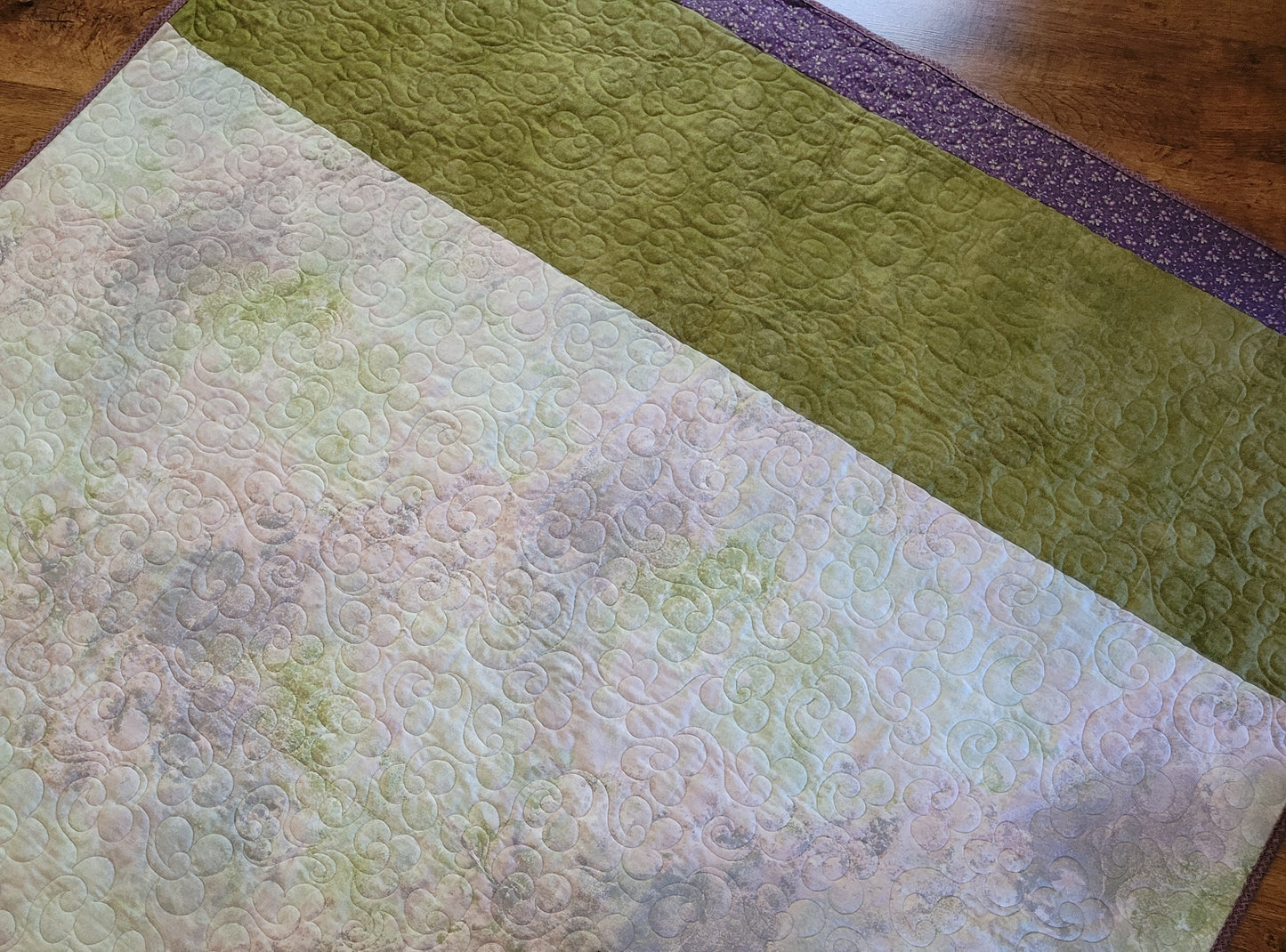 A close-up of the quilted throw blanket back, featuring a large pale blush and mint area with swirling stitched patterns, a wide moss green band, and a narrow purple patterned edge, laid on a wooden floor.