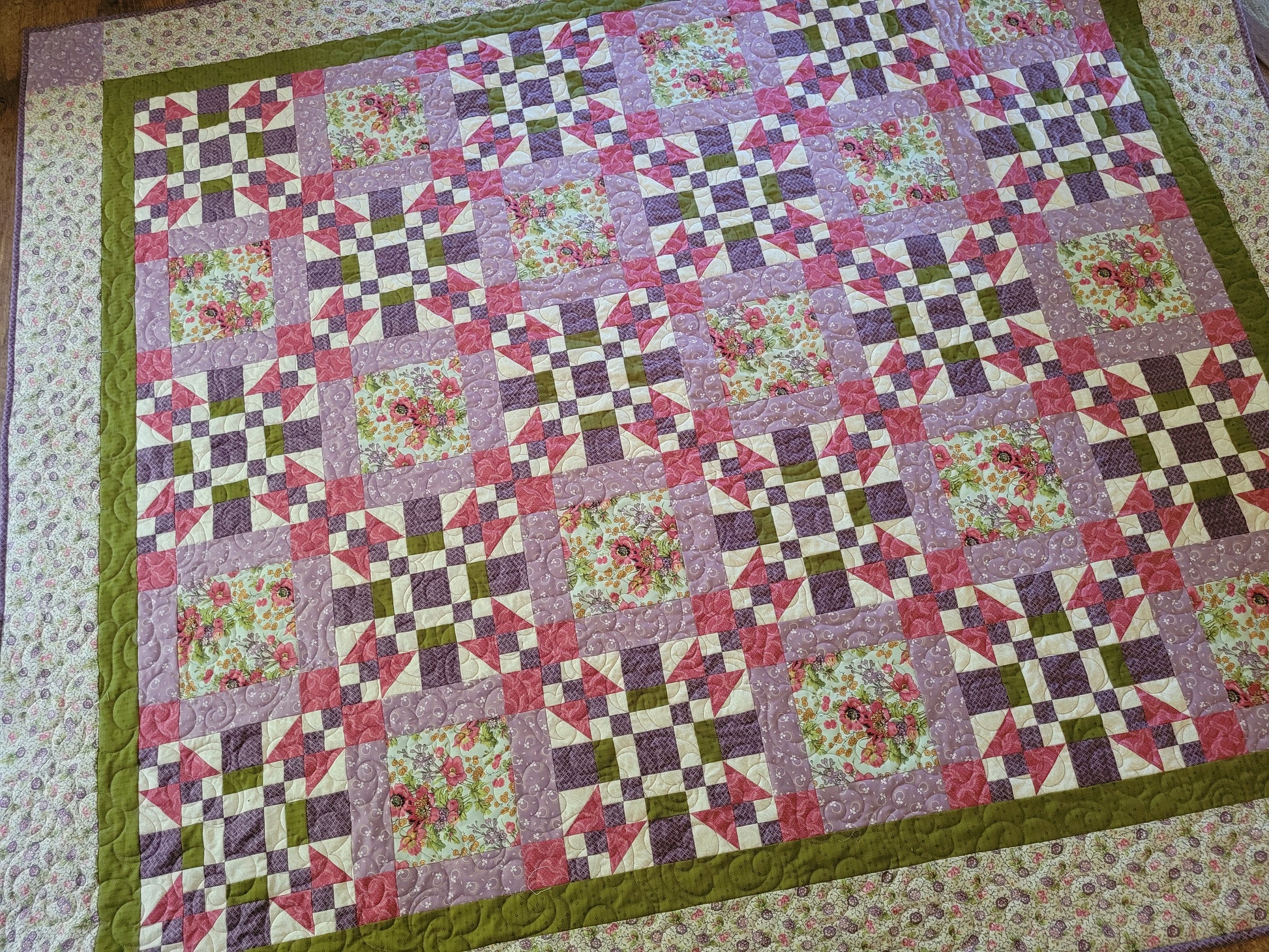 A handmade quilted blanket with a geometric and floral pattern, featuring pink, purple, green, and cream blocks, finished with an inner green border and a cream floral outter border.