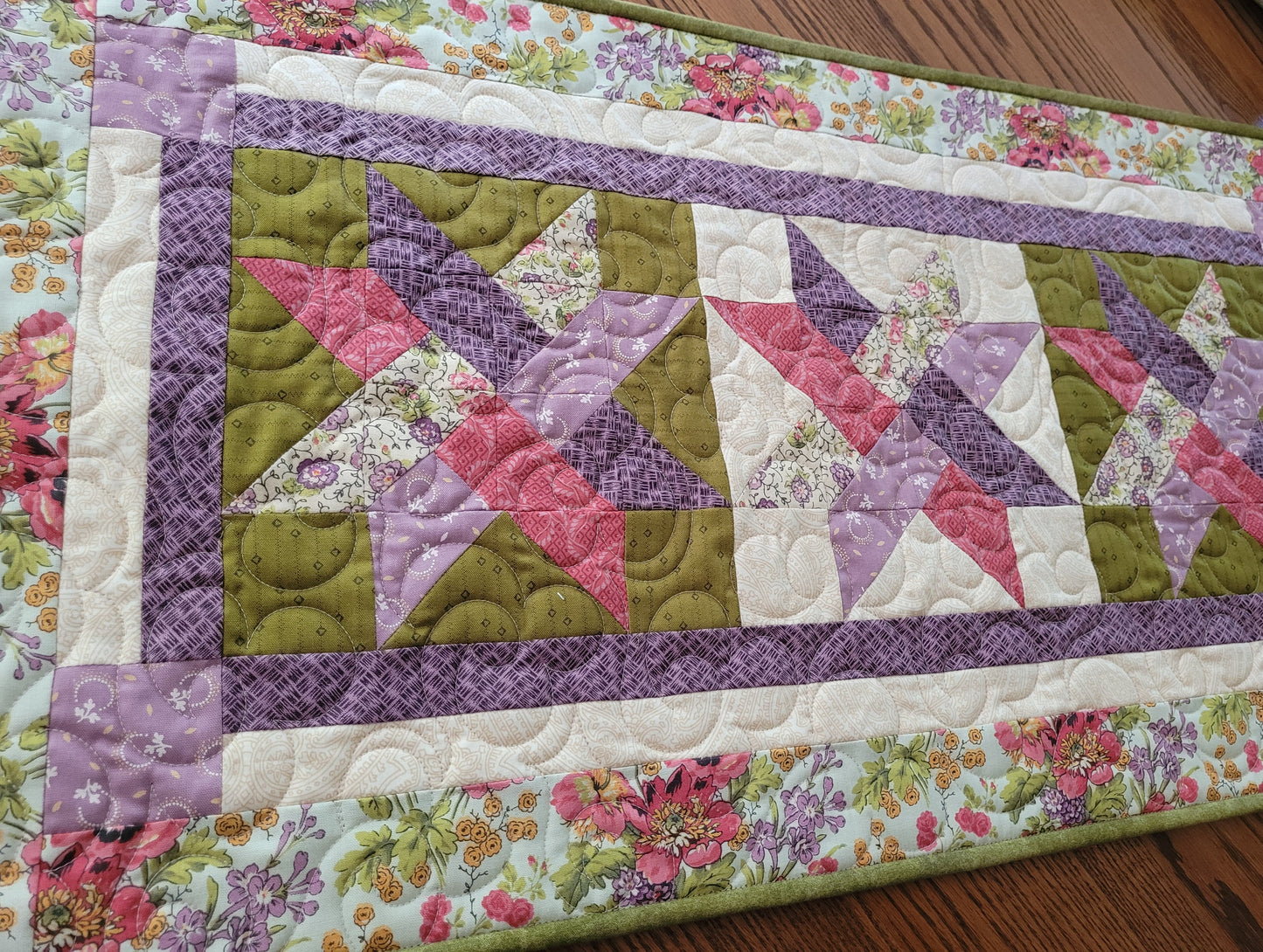 A handmade quilted table runner featuring a purple and mossy green woven star pattern with a floral printed border, laid flat on a wooden table.