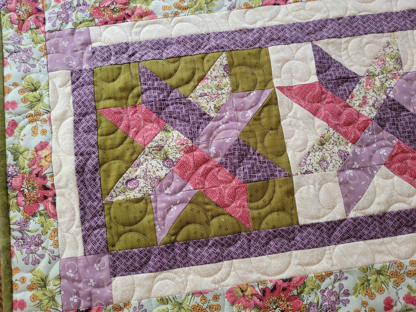 A close-up of a quilted table runner featuring a geometric woven star pattern in purple, moss green, and pink floral prints, bordered by a purple patterned strip and a floral border.