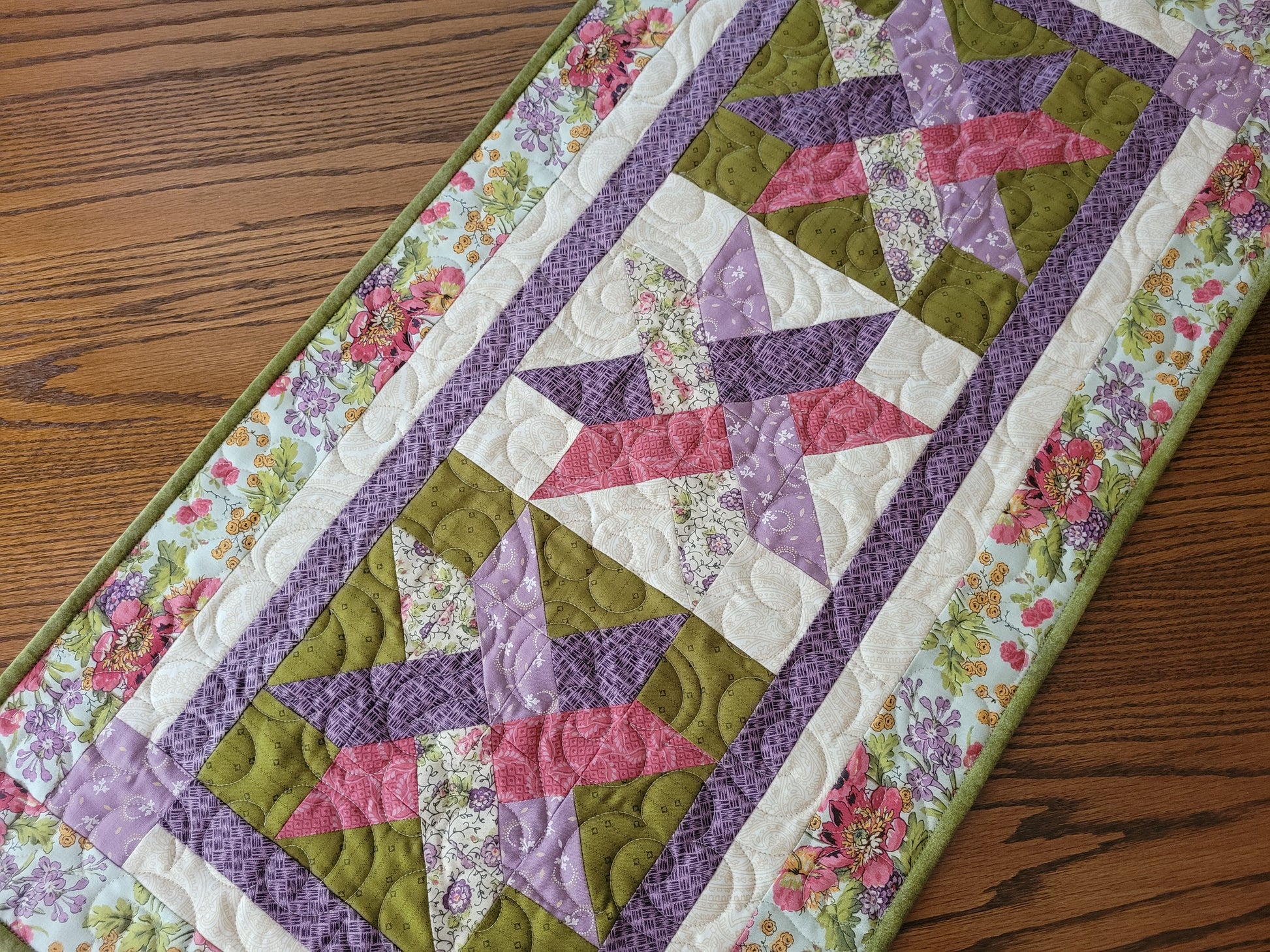 A close-up of a quilted table runner laid diagonally on a wooden table. The runner features a woven star pattern in purple, pink and mossy green, bordered by a coordinating floral print.