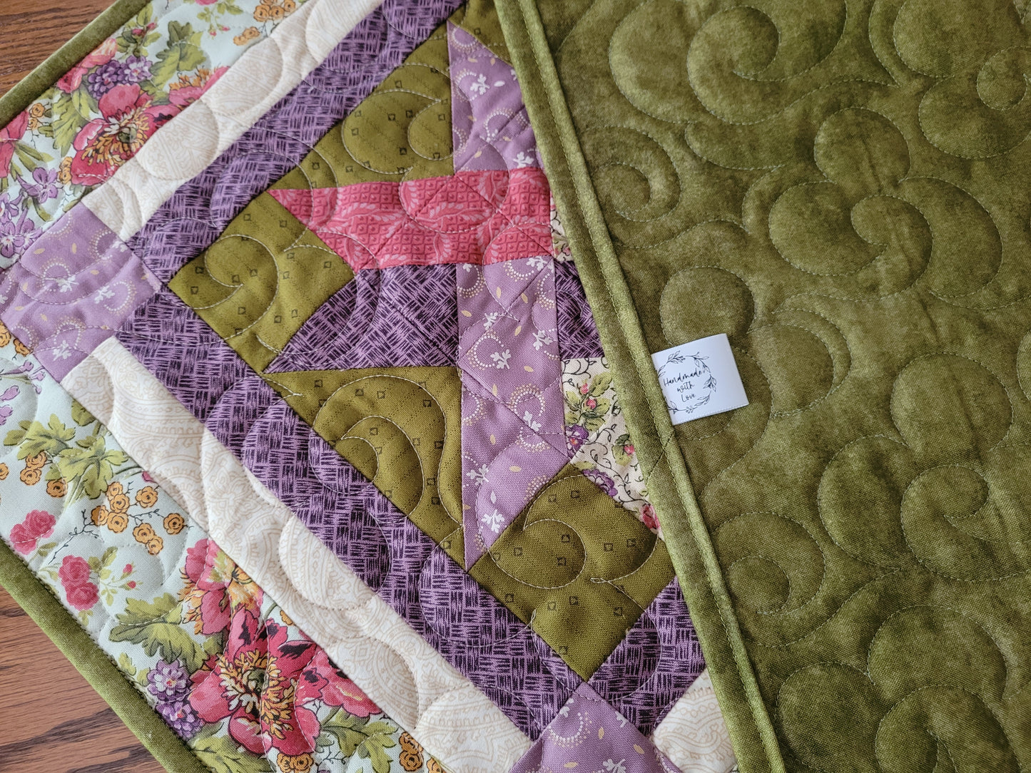 A close-up of the quilted table runner, showing a geometric patchwork design with moss green, lavender purple, pink, and floral cream sections. The right side features a solid moss green quilted backing with a stitched floral pattern.