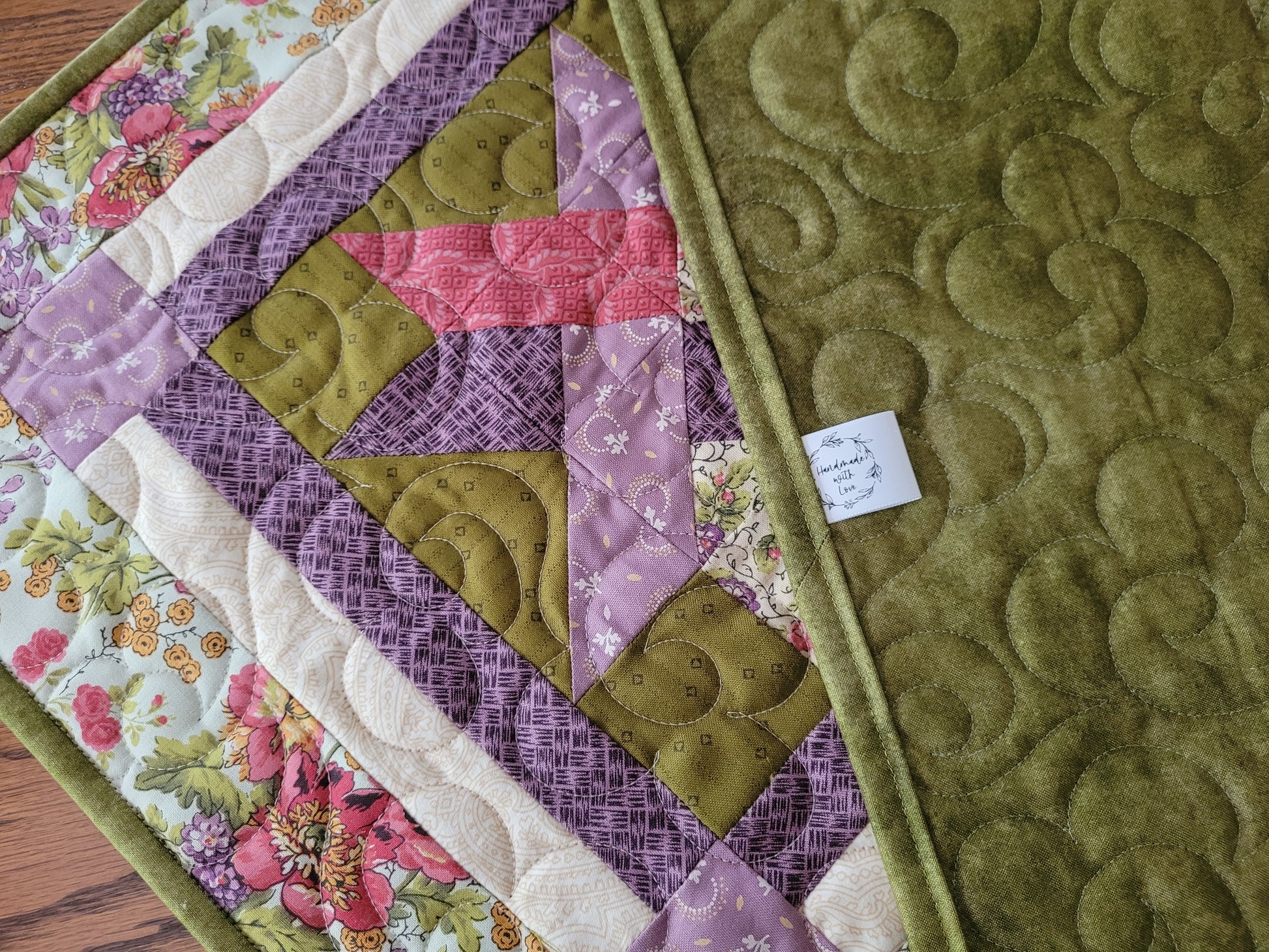 A close-up of the quilted table runner, showing a geometric patchwork design with moss green, lavender purple, pink, and floral cream sections. The right side features a solid moss green quilted backing with a stitched floral pattern.