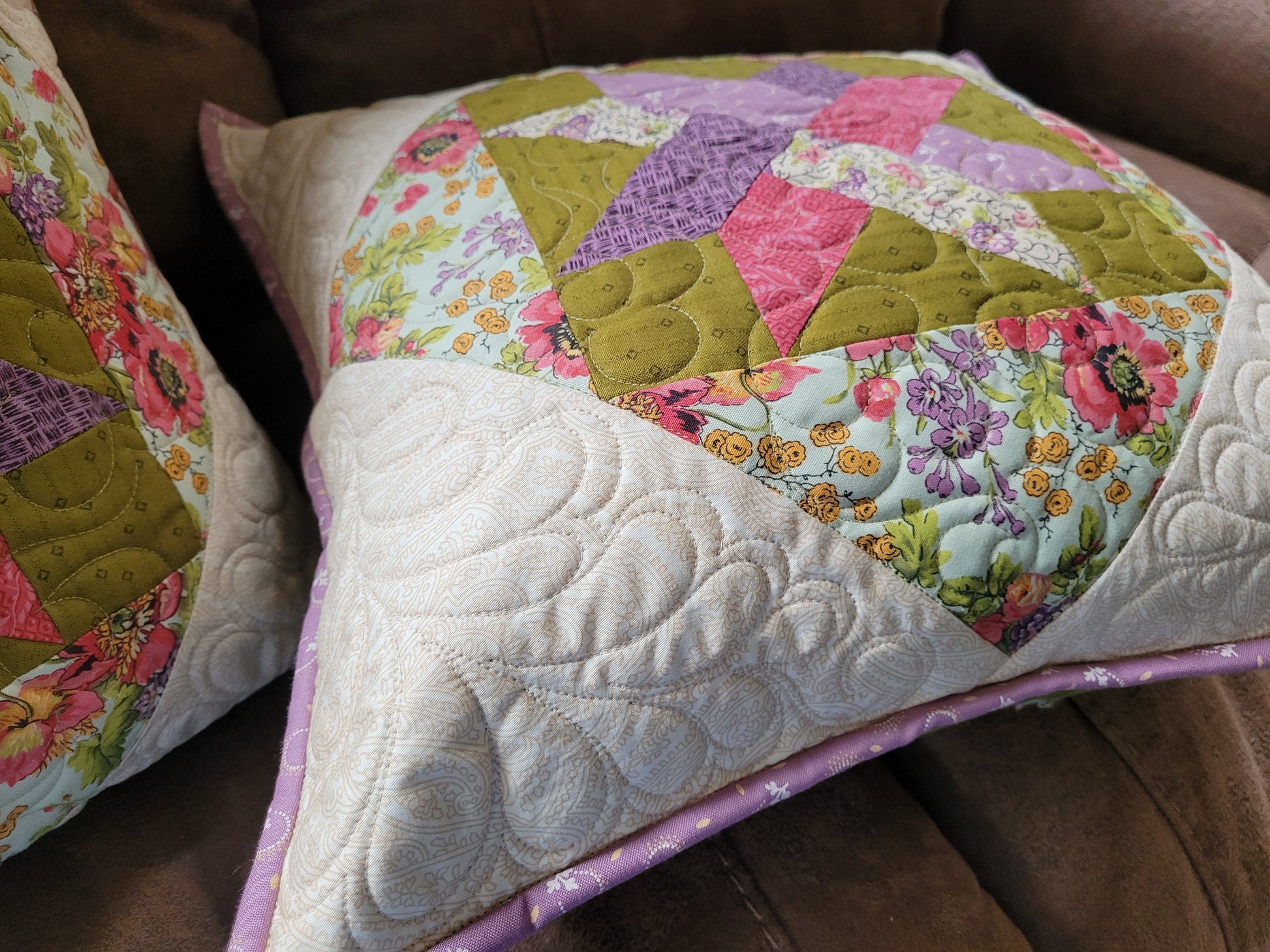 A close-up side angle view of a handmade quilted throw pillow with a patchwork design featuring moss green, pink, purple, and floral patterned patchwork, resting on a brown leather couch next to another pillow.