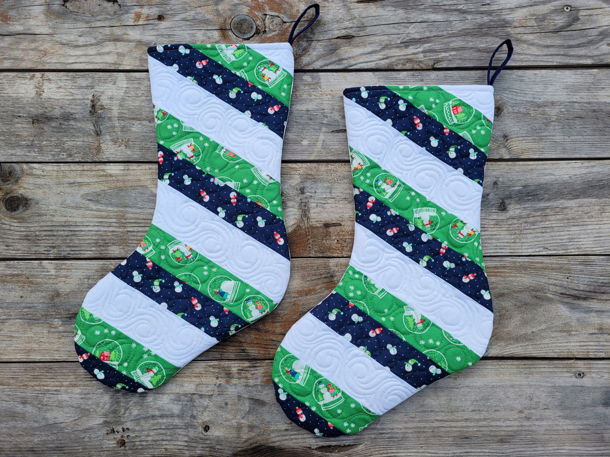 Two handmade Christmas stockings with green, white, and navy blue patterned stripes, displayed on a rustic wooden surface.