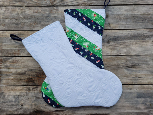 Two handmade quilted Christmas stockings laying atop each other, displayed on a rustic wooden plank background. Bottom one shows the stocking front in navy, green and white stripes and the stocking on top is showing the back with custom stitching.