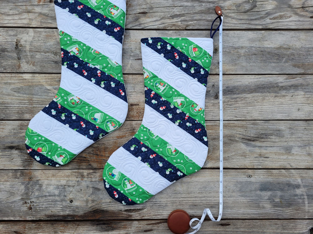 Two handmade Christmas stockings with a striped pattern of green, white, and navy blue fabrics featuring holiday motifs, displayed on a rustic wooden background. A tape measure showing measurements is laying beside.