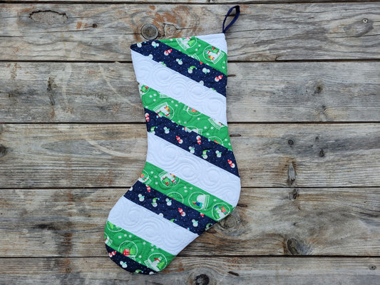 A handmade Christmas stocking made from striped fabric panels in green, navy, and white, featuring holiday patterns and a hanging loop, displayed flat on a rustic wooden plank background.