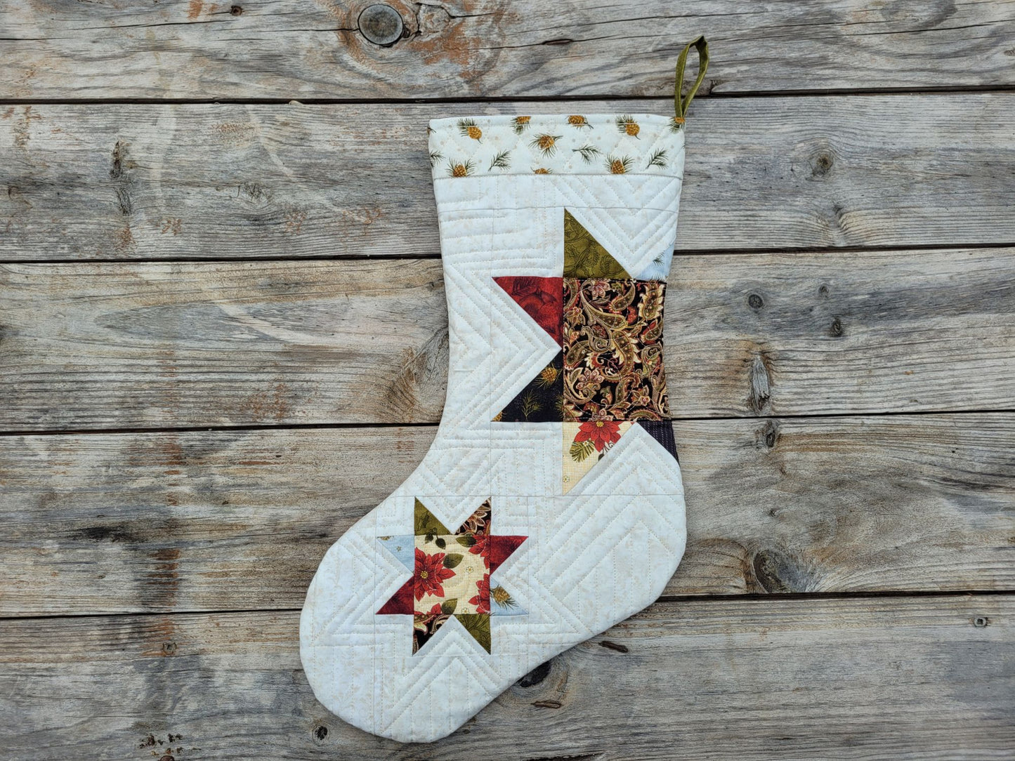 A handmade quilted Christmas stocking with a rustic patchwork design, featuring a white body, floral star patterns, and a green hanging loop, displayed on a weathered wooden plank background.