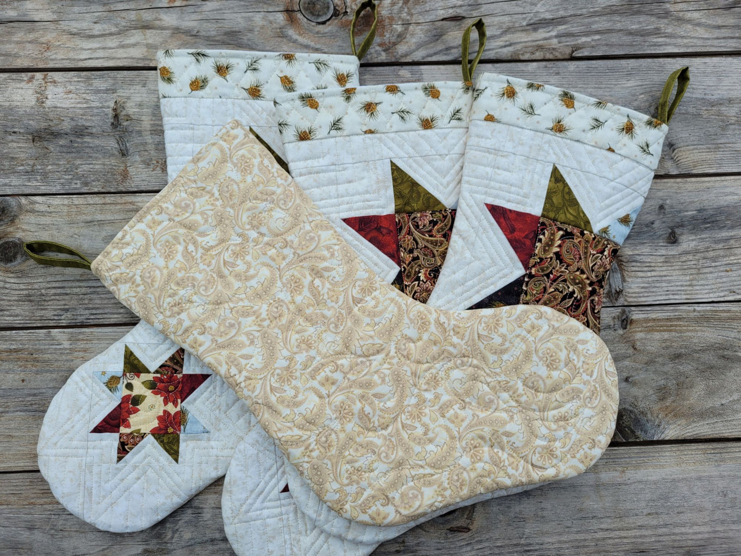 Quilted Christmas stockings arranged on a rustic wooden surface. One stocking is layed across the others with the back showing, which is a cream and beige damask pattern fabric. Stocking fronts feature star and floral patchwork accents in red, green, black and brown, and each has an olive green hanging loop.