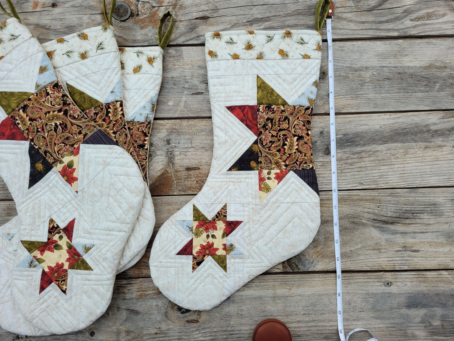 Handmade quilted Christmas stockings with star and floral patchwork designs, laid flat on a rustic wooden surface next to a white measuring tape.