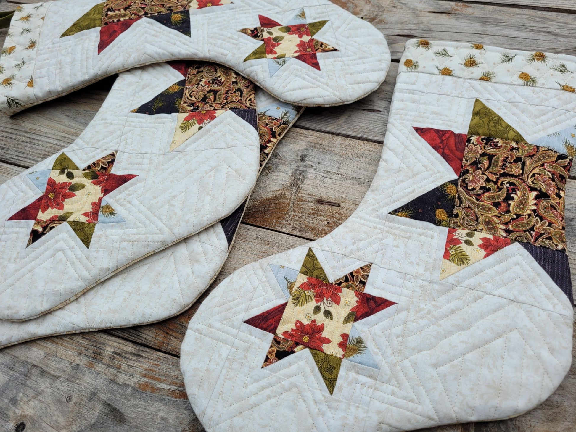 Four quilted Christmas stockings with star patterns, featuring a mix of cream, red, green, and brown fabrics, arranged on a rustic wooden surface.