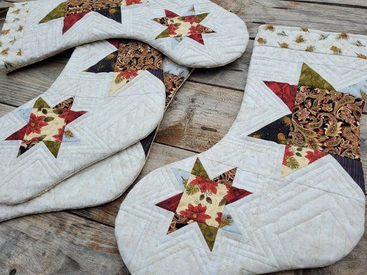 Four quilted Christmas stockings with star patterns, featuring a mix of cream, red, green, and brown fabrics, arranged on a rustic wooden surface.