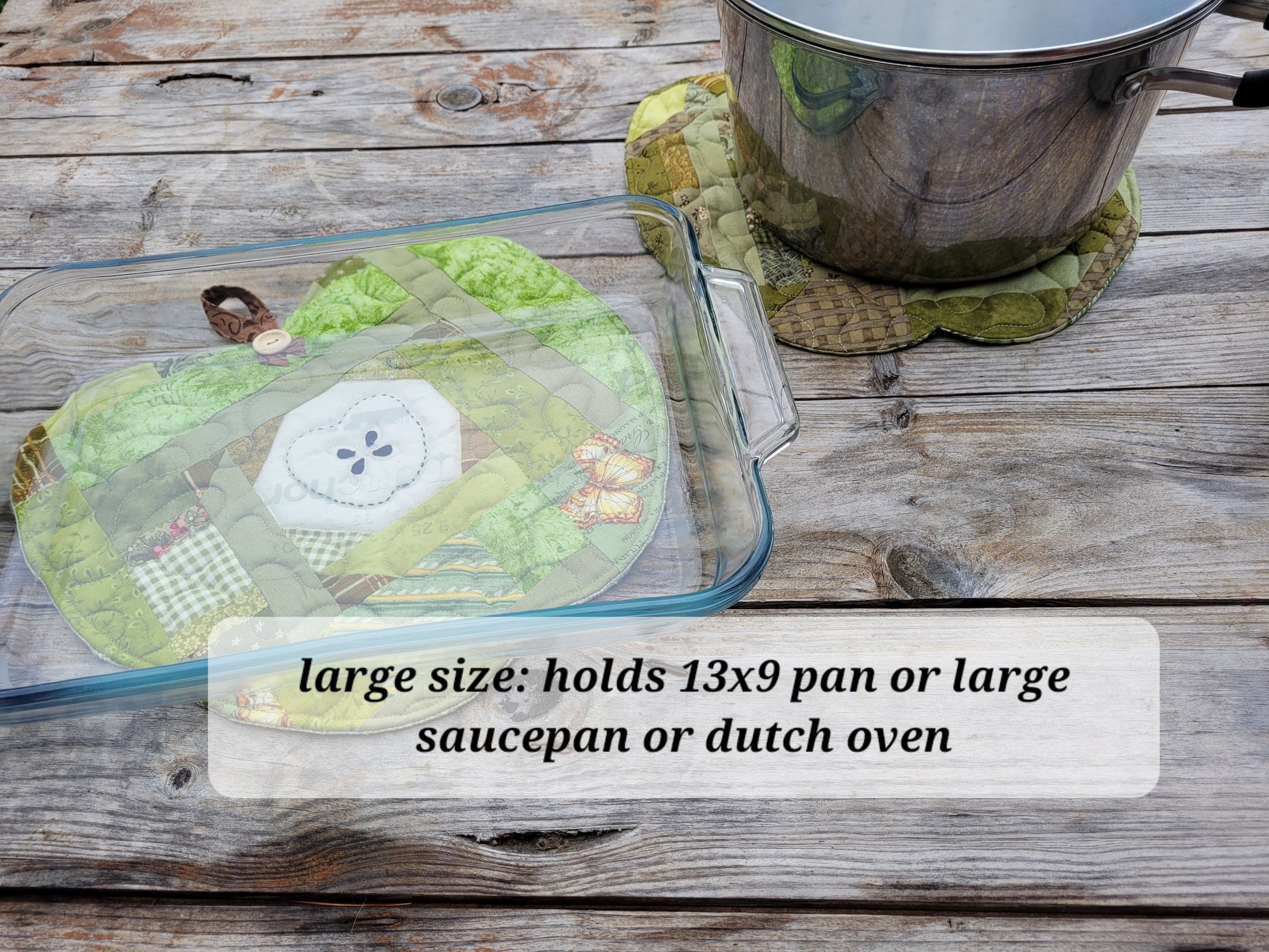 A glass pan and stainless steel pot are sitting on two green apple shaped quilts on a rustic gray wooden table. A translucent text box says, 'large size holds 13x9 pan or large saucepan or dutch oven'.