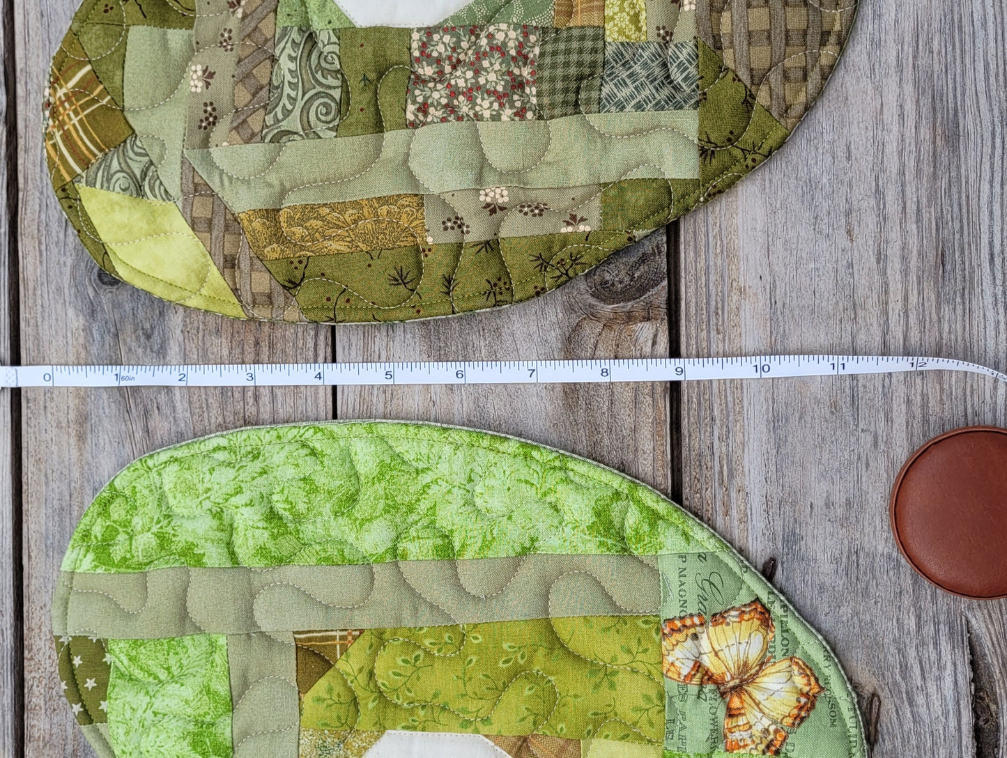 A white measuring tape is laying between two partially visable green apple mini quilts on a rustic gray wooden table 