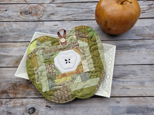 A quilted fabric potholder shaped like an apple, featuring a patchwork design in various shades of green and beige with a white stitched apple center, displayed on a white lace plate over a rustic gray wooden table with a gourd in the background.