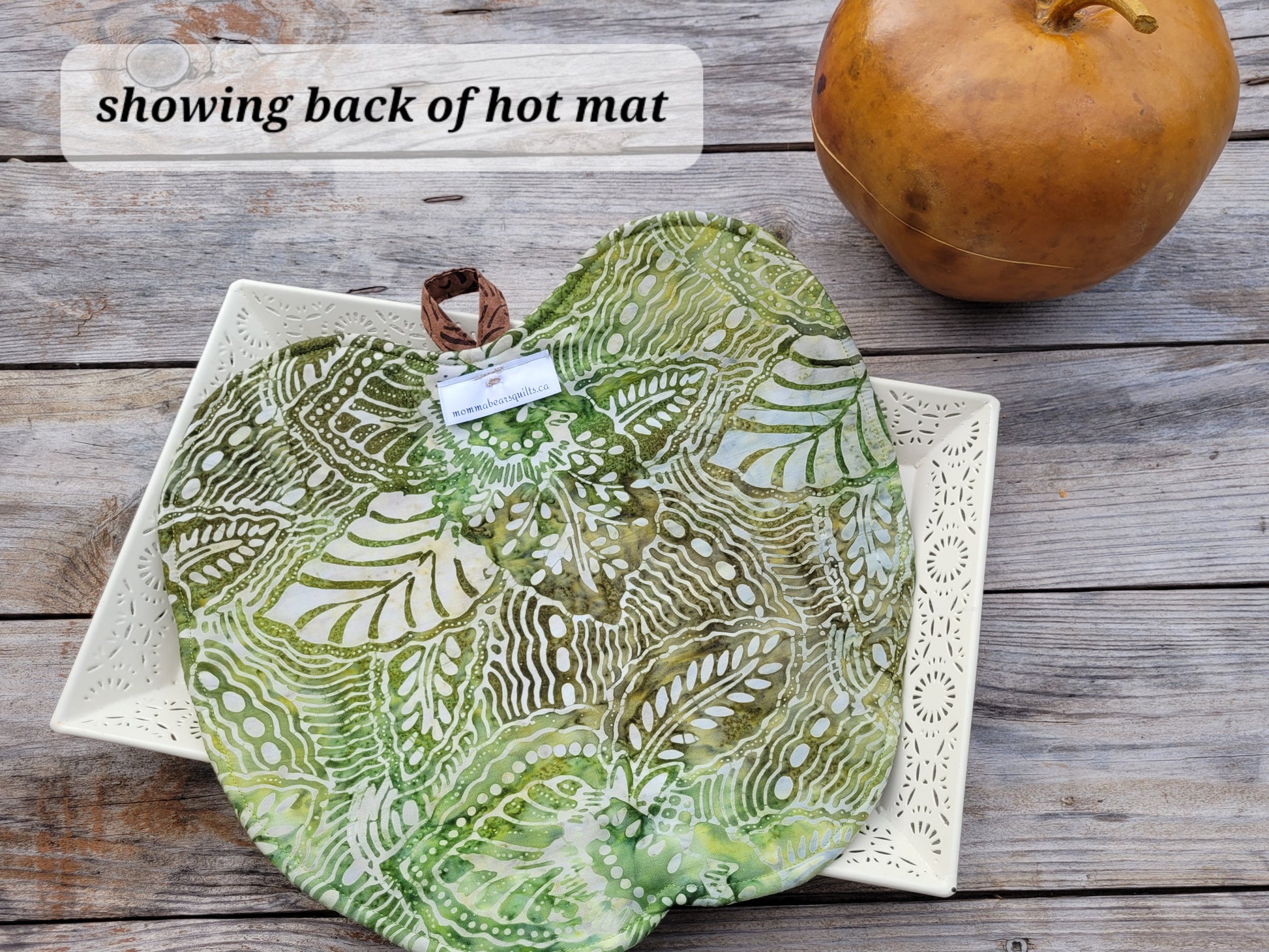 A green apple shaped quilt is laying on a white decorative tray atop a gray wooden suface with an apple shaped gourd in the background. Translucent text box says 'showing back of hot mat'.