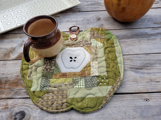 A quilted apple patchwork trivet or placemat in muted green, beige, and brown tones, featuring a central white stitched apple motif. A brown ceramic mug sits on top, and the item is photographed on a rustic wooden surface with other home decor props partially visible.
