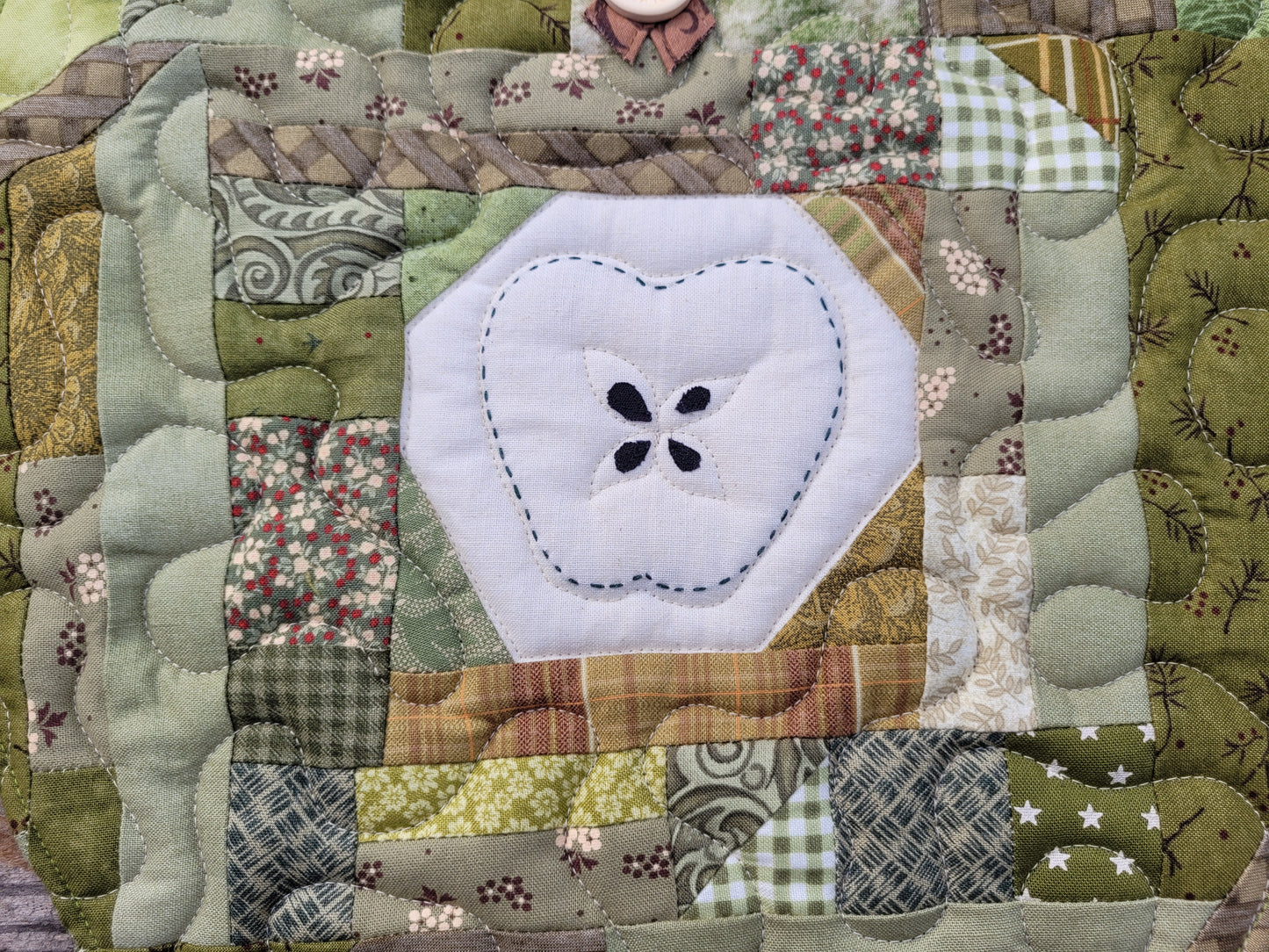 A close-up of a green scrappy patchwork quilt with a central muslin patch with a hand-stitched apple outline around four appliqued seeds. 