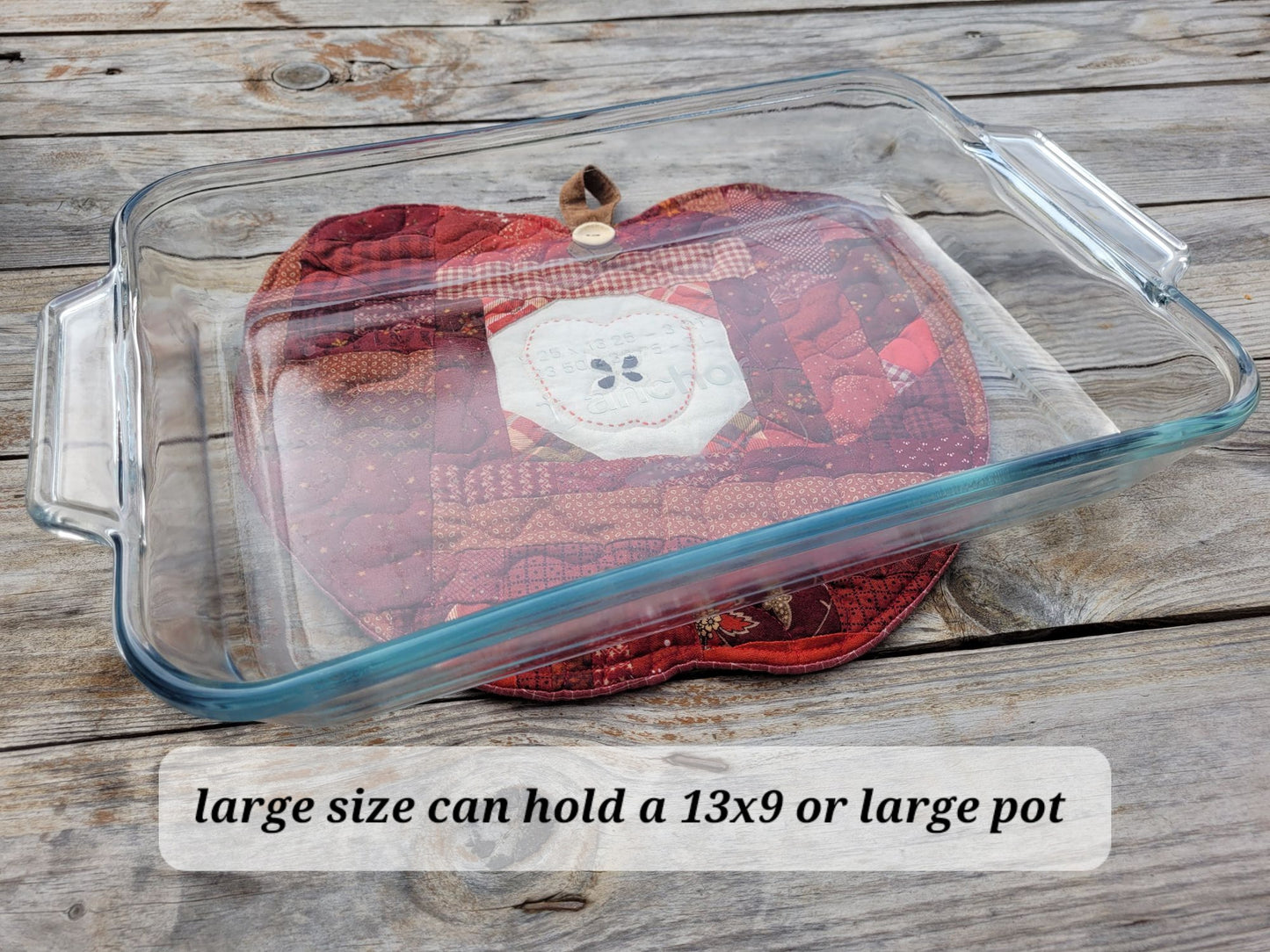 A large clear glass baking dish with handles, shown on a rustic wooden surface with a red quilted apple mat underneath. Text at the bottom reads, 'large size can hold a 13x9 or large pot'.